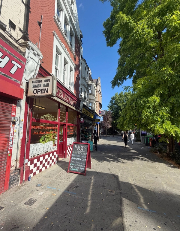 Street-level exterior view of a red shopfront cafe set on a sunlit South London high street pavement in Tulse Hill, Love Locations SE27.