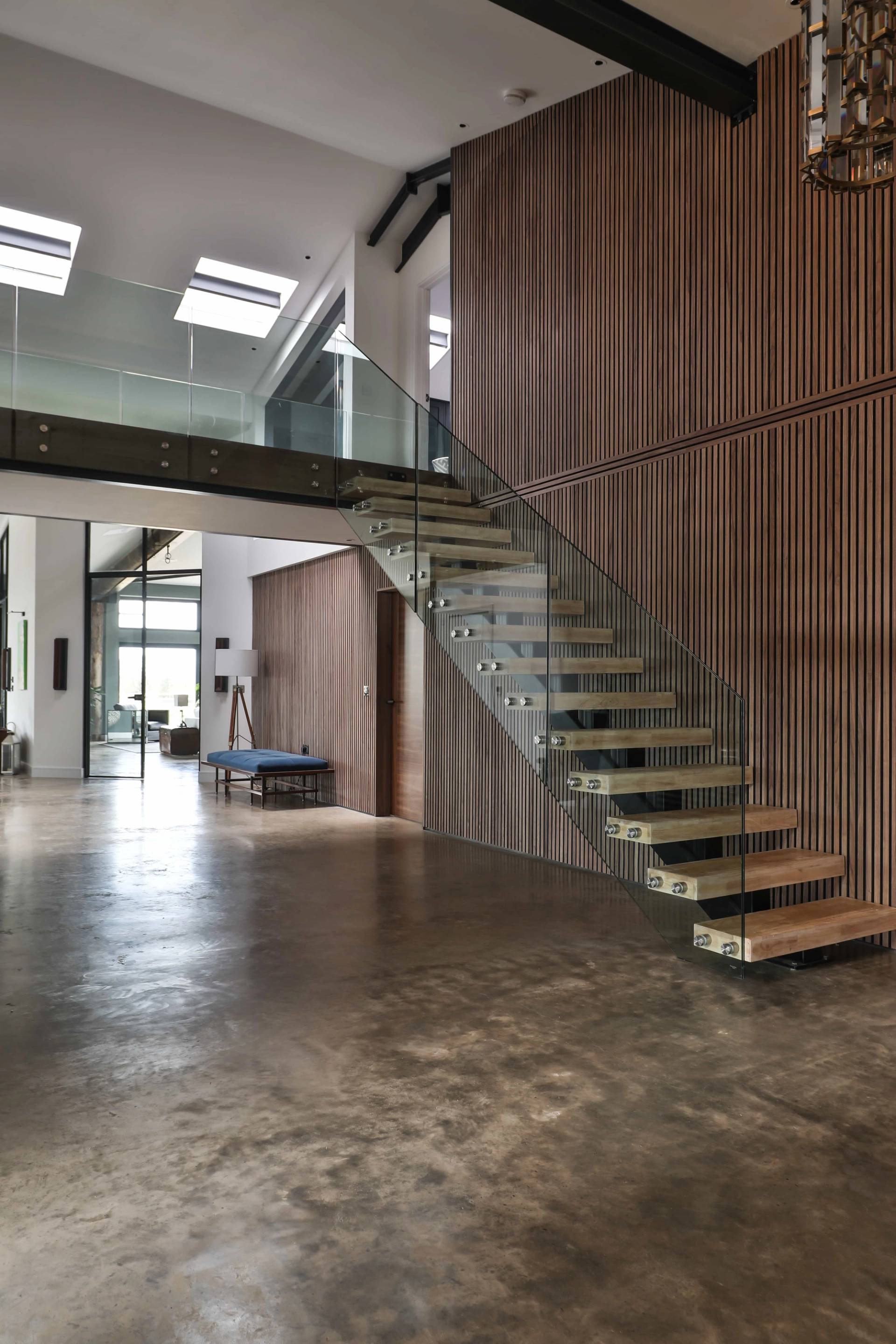 Under-stair view of a floating wooden staircase with glass balustrades in a double-height barn hall, Love Locations CV23.