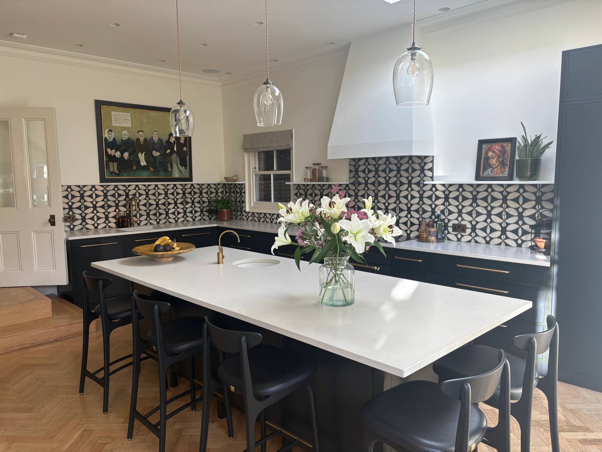 Contemporary kitchen island with white countertop, black bar stools, vase of lilies, and a striking black-and-white patterned tile splashback in Twickenham TW1 via Love Locations.