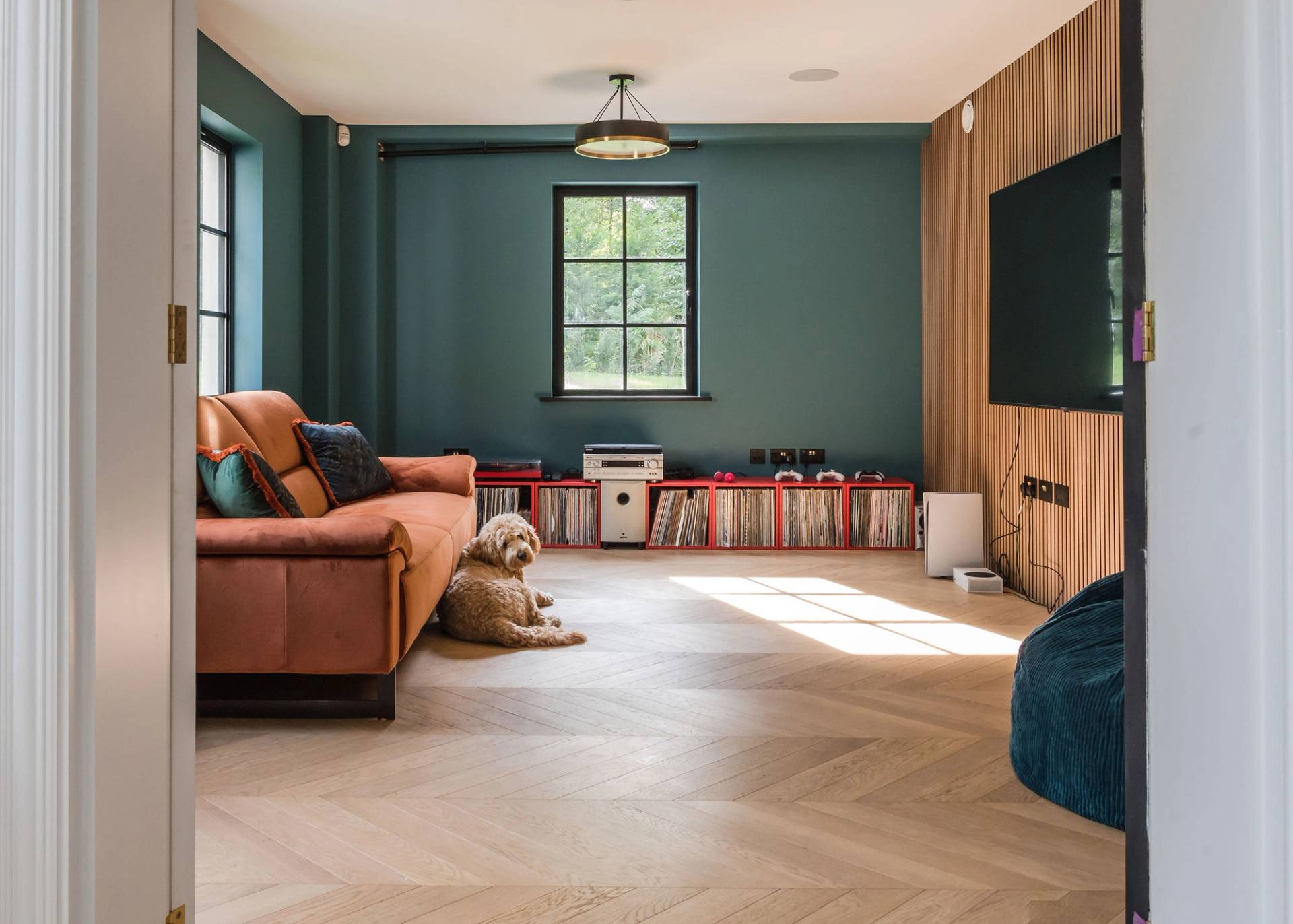 Cozy family cinema room with a rust-orange sectional sofa, oak panelled TV wall, green accent wall, and parquet floors, Love Locations GU3.