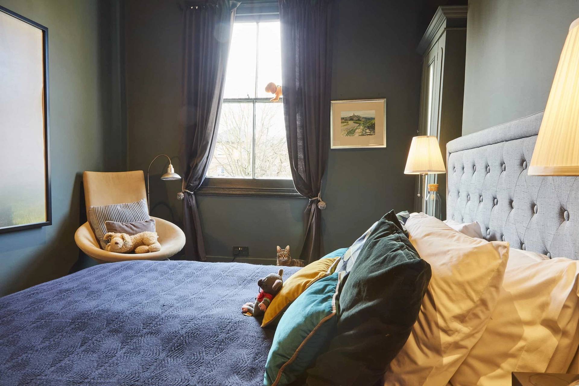 Dark teal bedroom corner with dark window drapes, mid-century armchair, and soft lighting, Love Locations N5.
