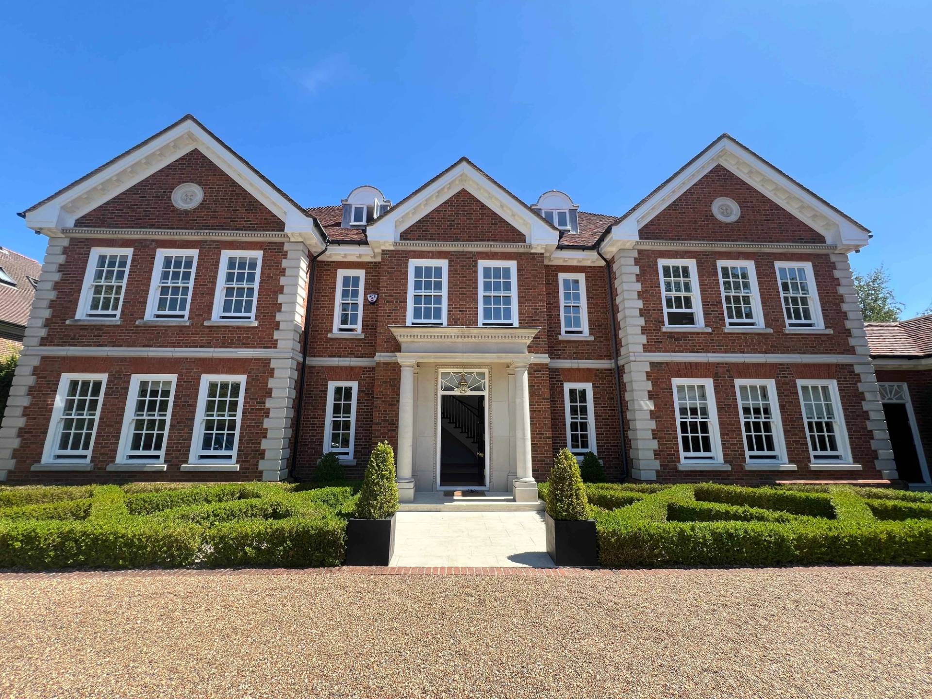 Double-fronted red brick mansion front elevation in BR7 with gravel driveway, stone portico, and tiered fountain, Love Locations Bromley.