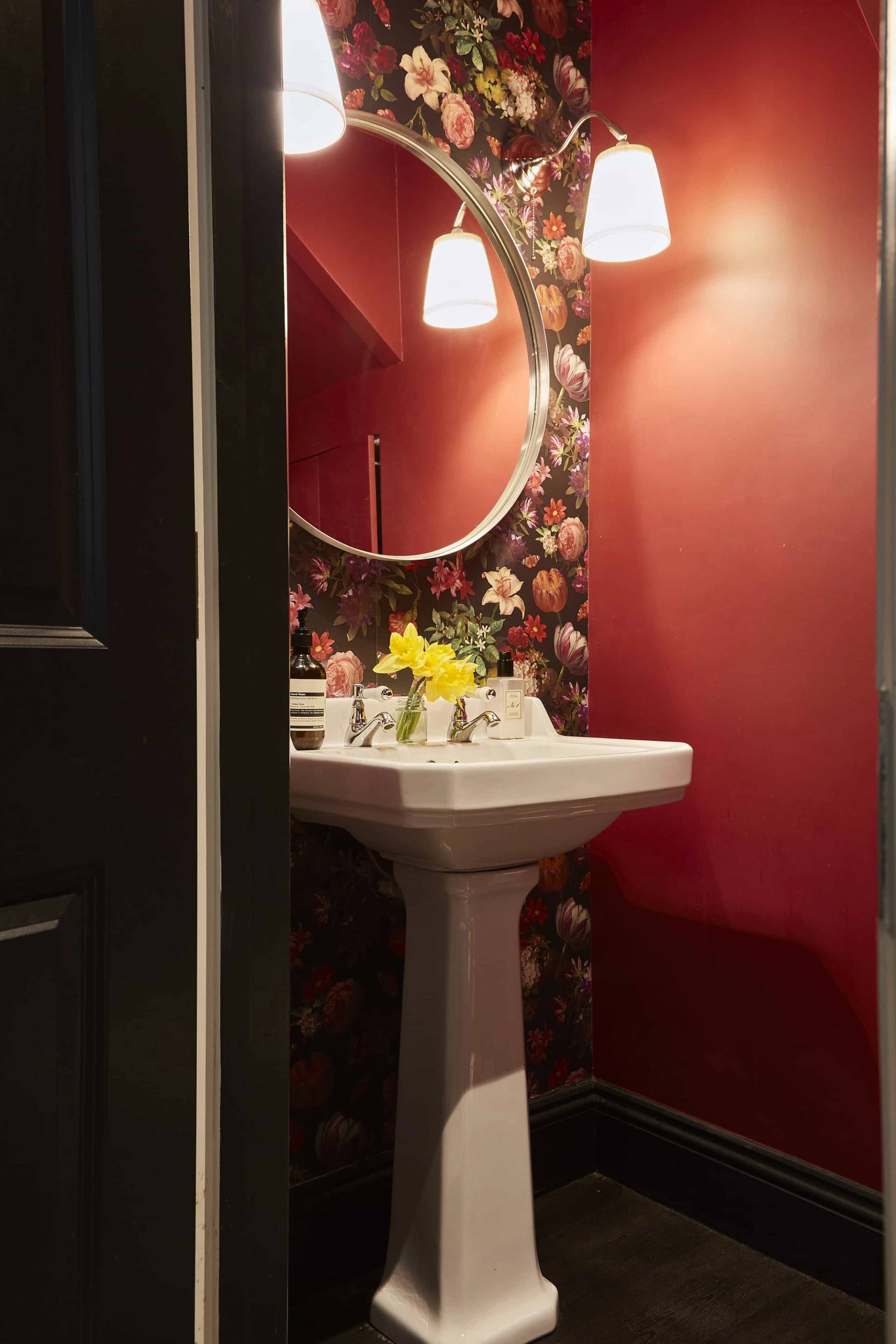 Powder room bathroom featuring dramatic deep red painted walls, floral wallpaper, white pedestal sink, and round mirror, Love Locations.