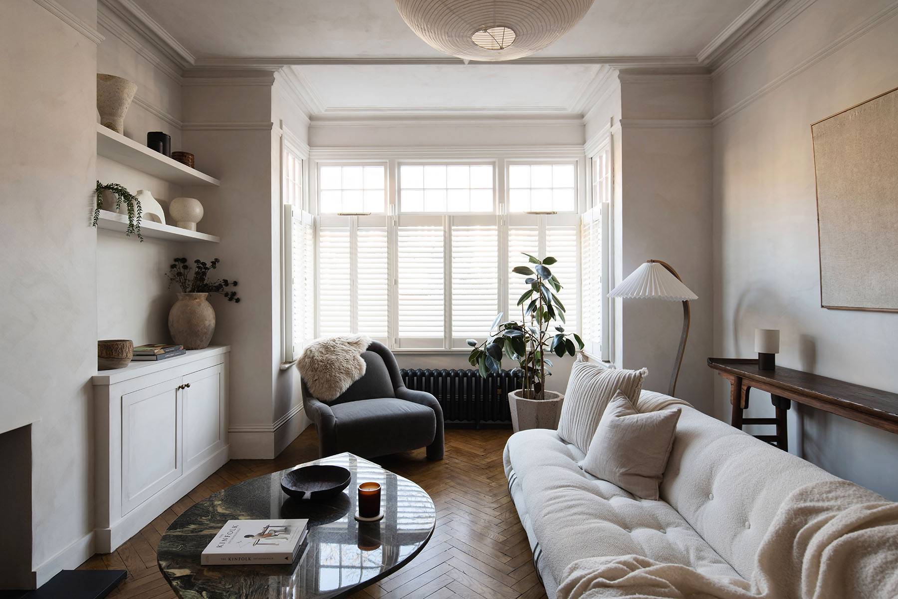 Wabi-sabi lounge room featuring raw micro-cement walls, herringbone parquet floors, white sofa, and bay window shutters, Love Locations.