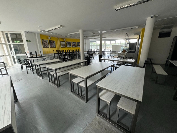 Love Locations modern school cafeteria hub Cockfosters: Clean eating area showing bench-style wooden dining tables under bright flourescent panel lights, configured for student lifestyle photography.
