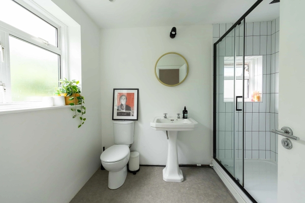 Love Locations modern bathroom location East London E7: Contemporary white washroom featuring a pedestal sink, round brass mirror, pop art frame, and grid glass walk-in shower, optimized for beauty ads.
