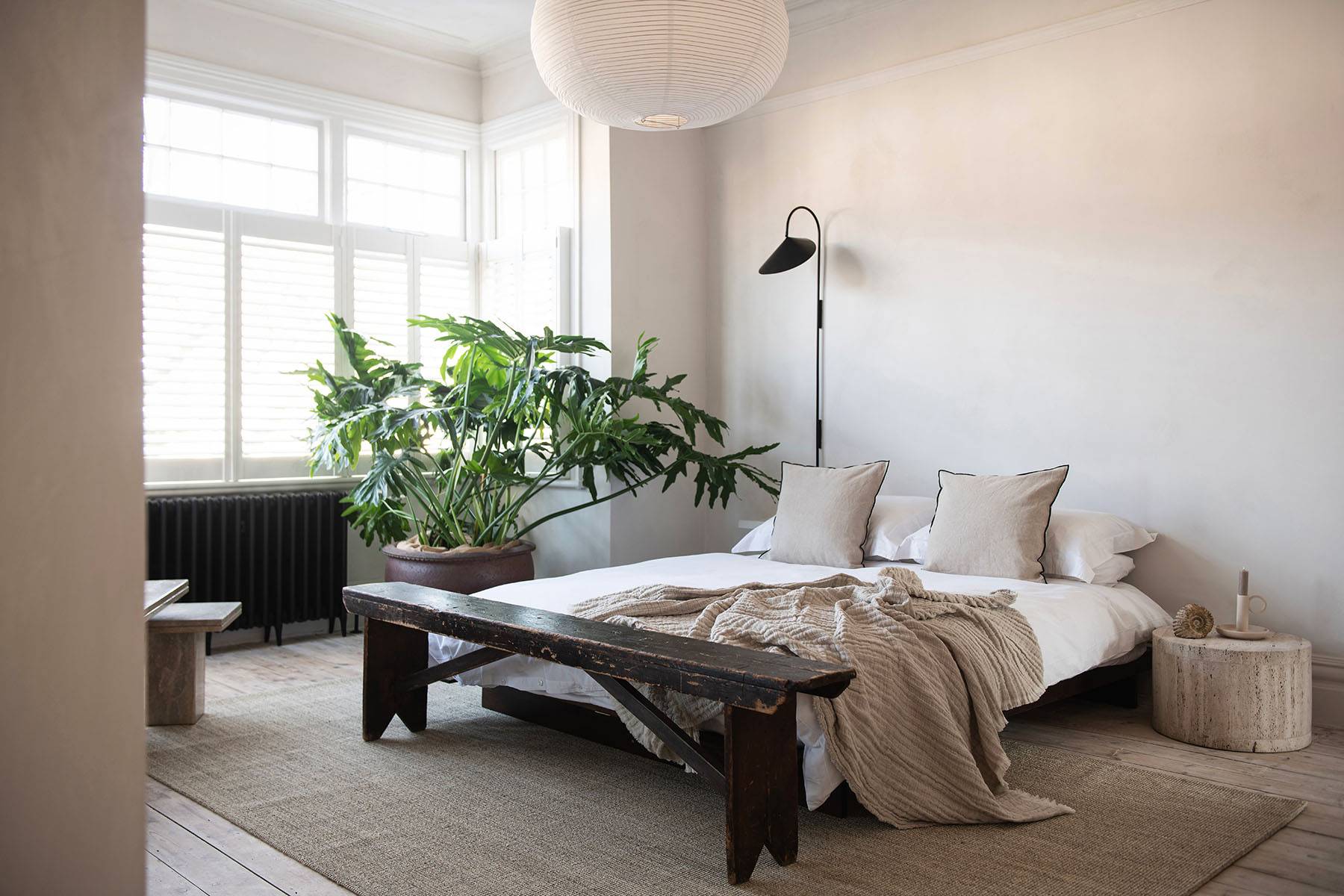 Light-filled bedroom with low wooden platform bed, white window shutters, and large leafy green potted plants, Love Locations SW16.
