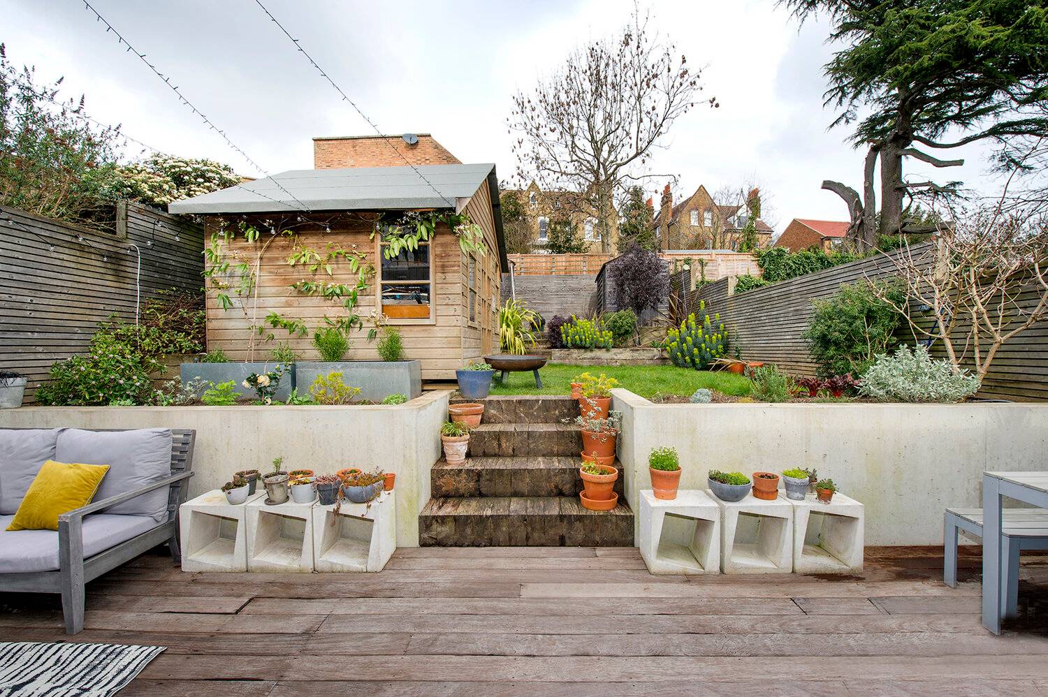 Landscaped garden patio in Forest Hill, SE23 featuring tiered timber decking, smooth concrete retaining walls, rustic wooden steps, potted terracotta plants, grey outdoor seating, and a standalone timber garden studio hut, Love Locations SE23.