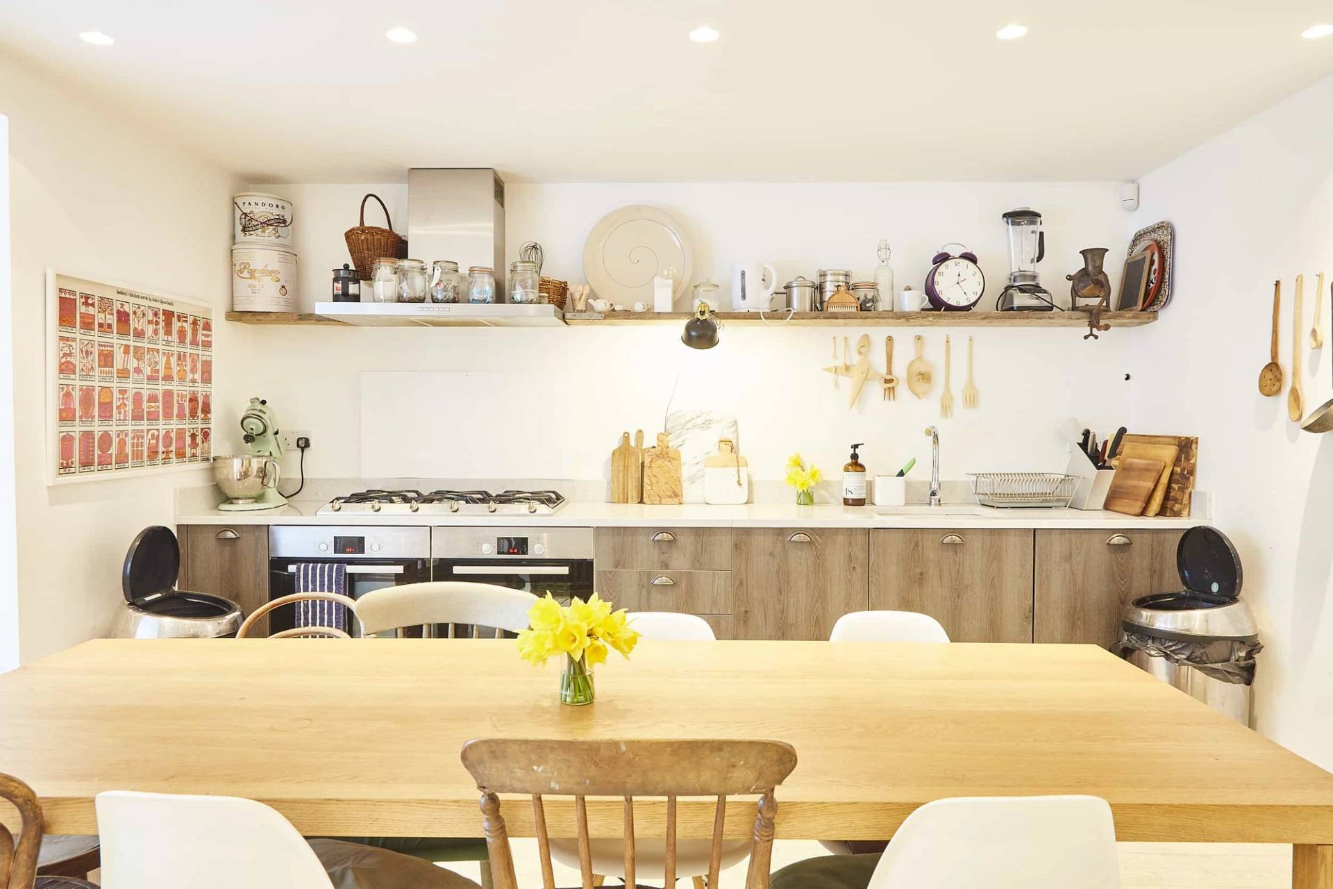 Long wooden dining table set with yellow flowers set before open timber wall shelves filled with copper kitchenware, Love Locations N5.
