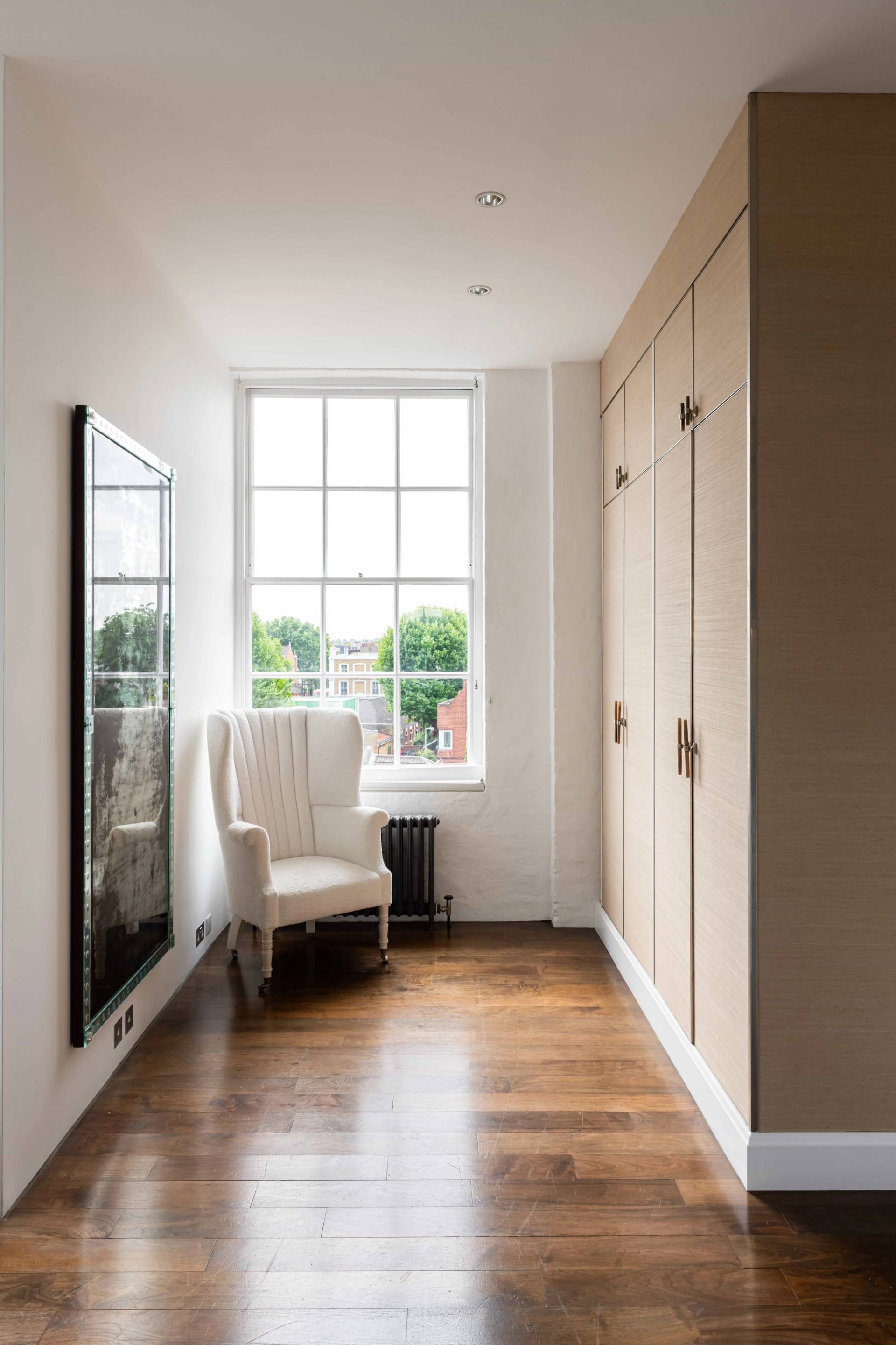 Dressing room corner with built-in beige oak wardrobes, white wingback armchair, black radiator, and sash window light, Love Locations.