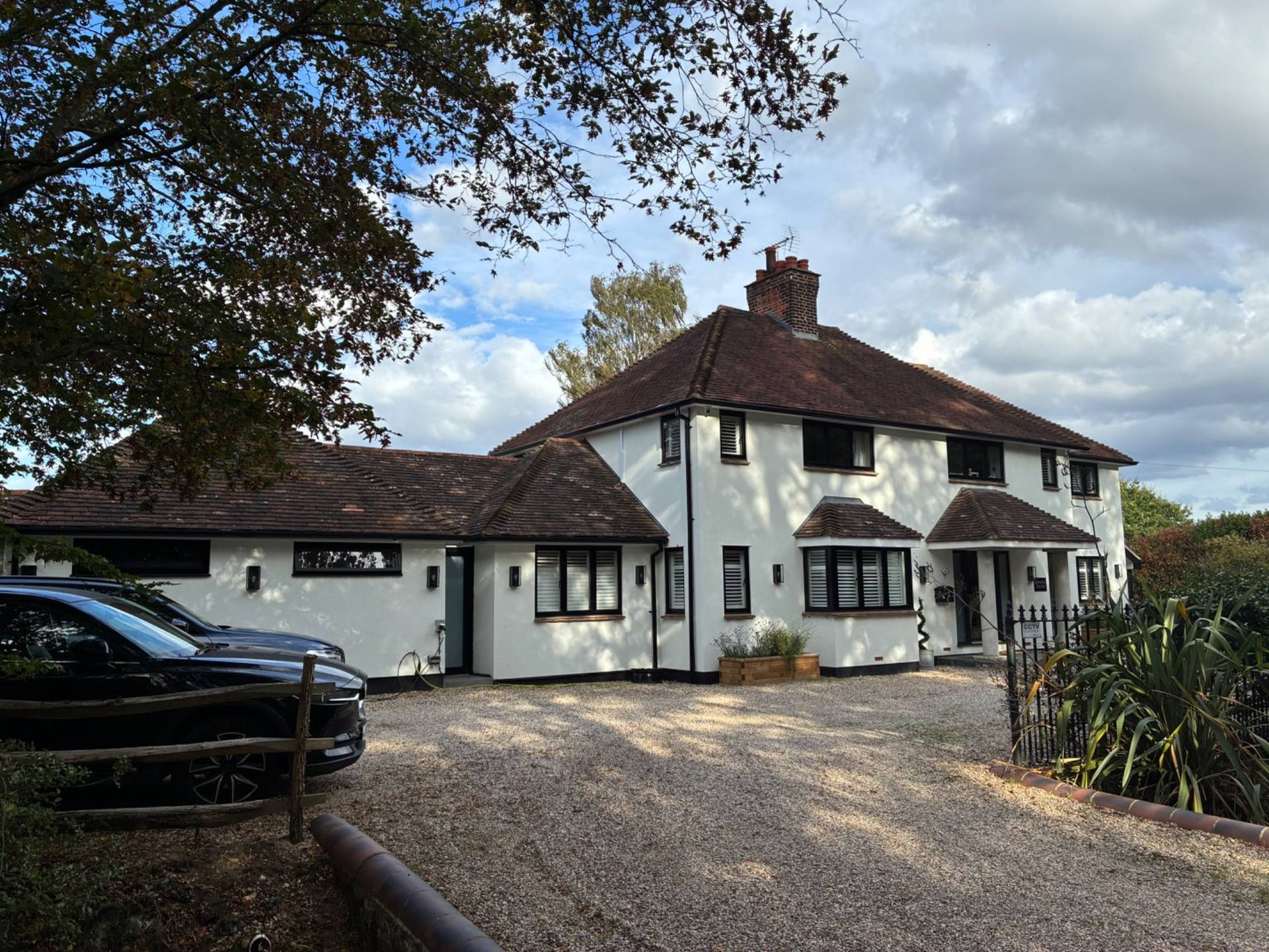Exterior front view of an exclusive private country house location in Essex CM4 by Love Locations, showing lush surrounding trees, security gates, and a spacious gravel driveway.