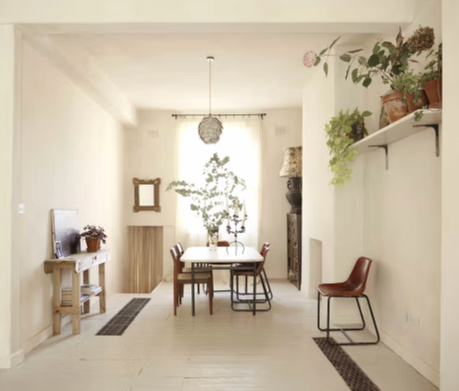 Minimalist dining room at Portobello location house featuring white-painted floorboards, a simple wooden dining table, vintage chairs, floating plant shelves, and abundant natural light.