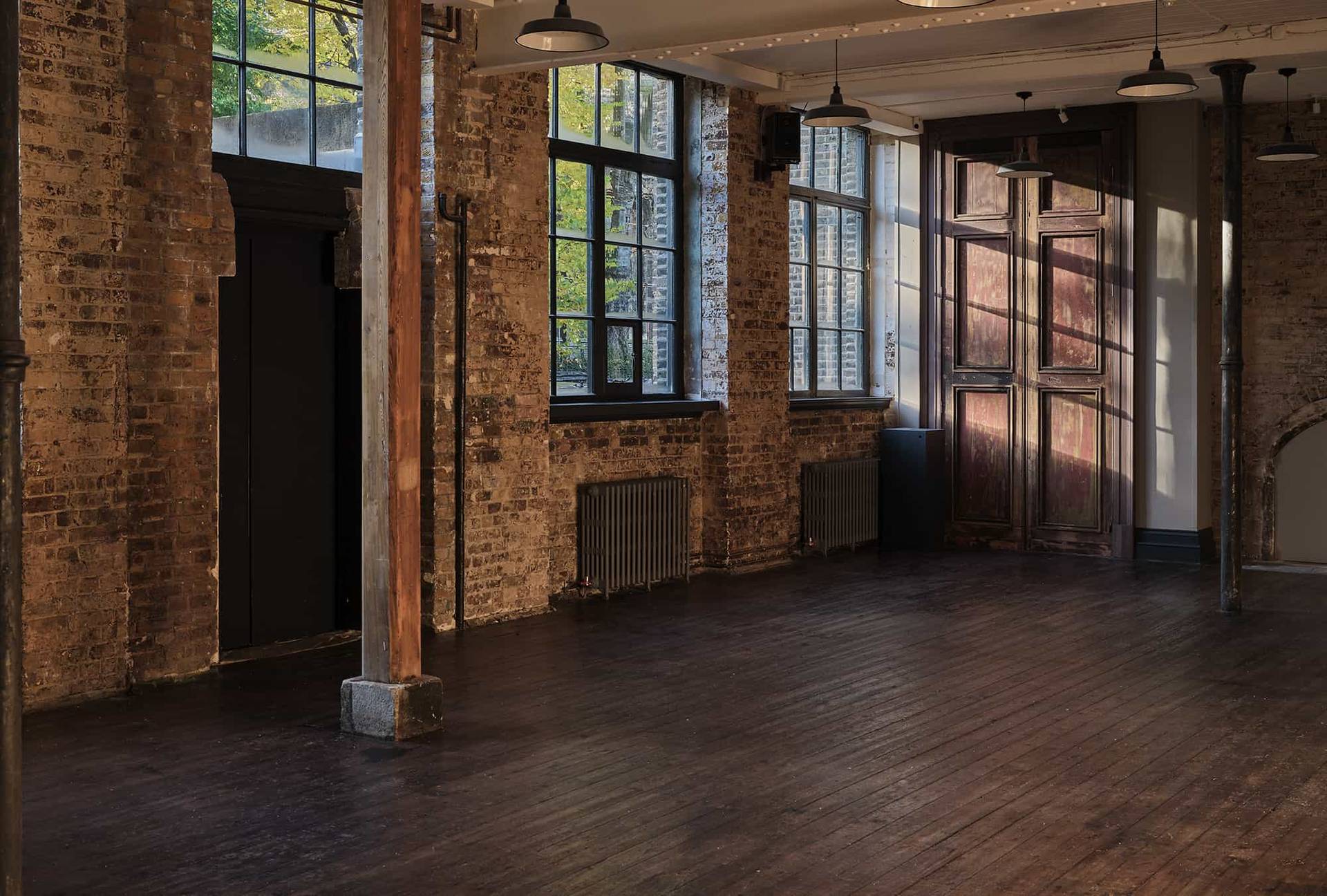 Corner perspective of an industrial brick venue with wooden columns, large windows, and dark floorboards, Love Locations E2.