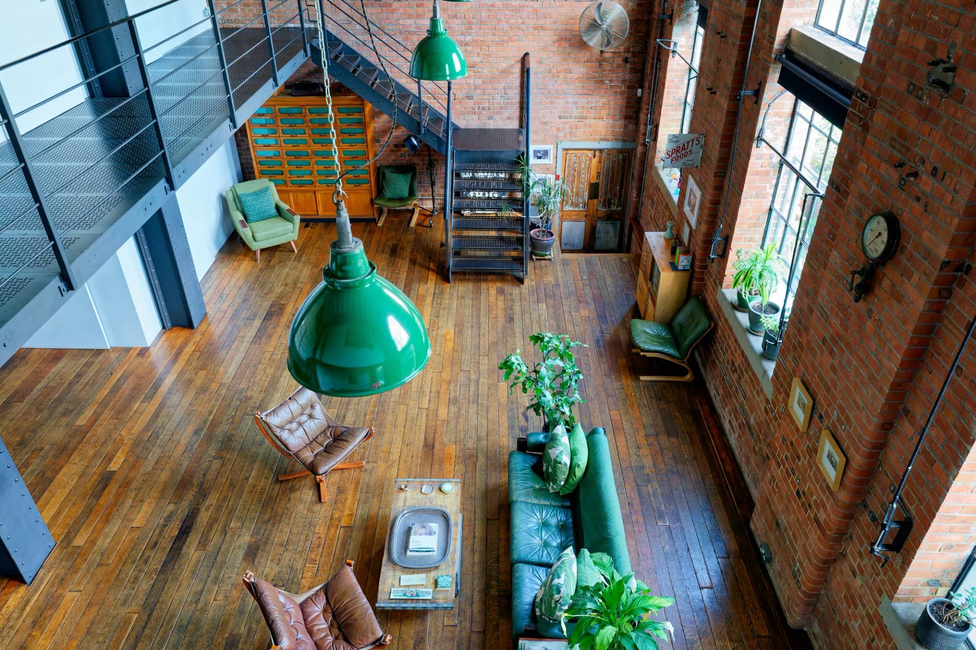 High-angle bird's-eye view looking down from a mezzanine onto a green velvet sofa, leather armchairs, and timber floors, Love Locations E14.