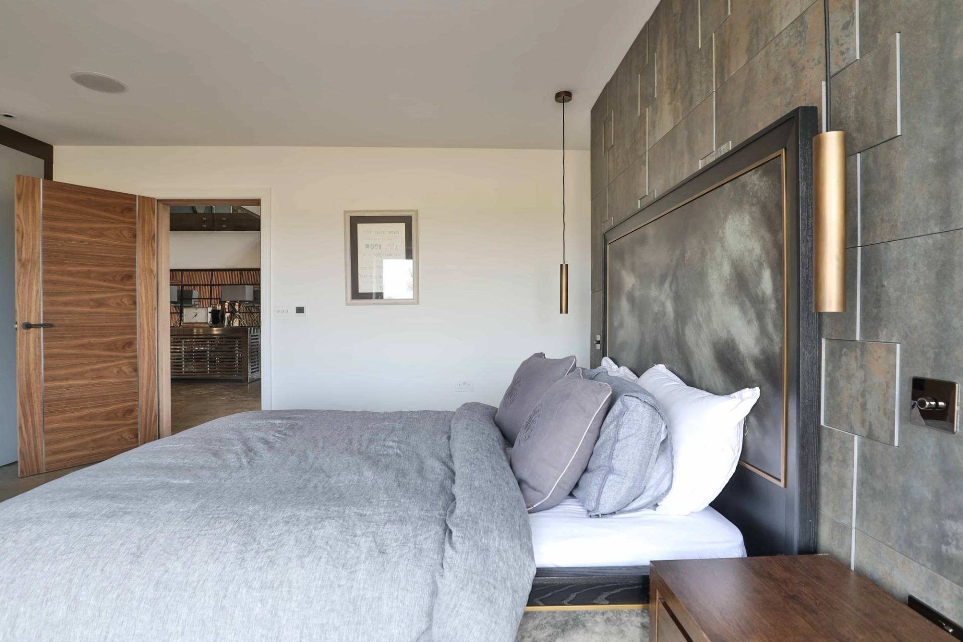 Side view of a master bed setup with stone-texture feature wall, pendant lighting, and grey linen, Love Locations CV23.