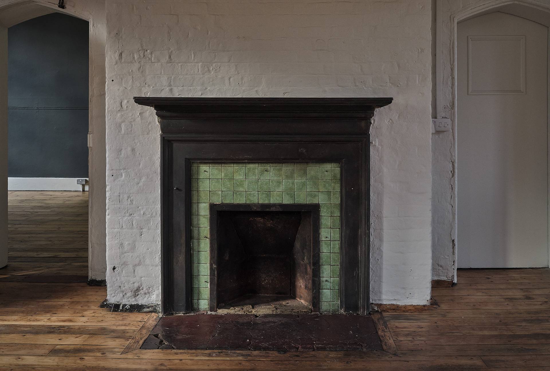 Period fireplace surround with green tiles and dark timber mantelpiece set against white brickwork walls, Love Locations E2.