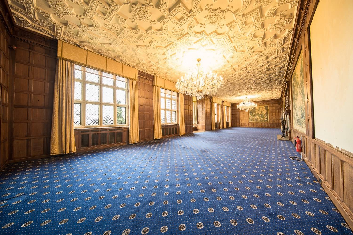 Spacious long gallery ballroom filming location in Hertfordshire AL9 through Love Locations, showcasing an intricate plasterwork ceiling, crystal chandeliers, blue carpet, and grand lattice windows.