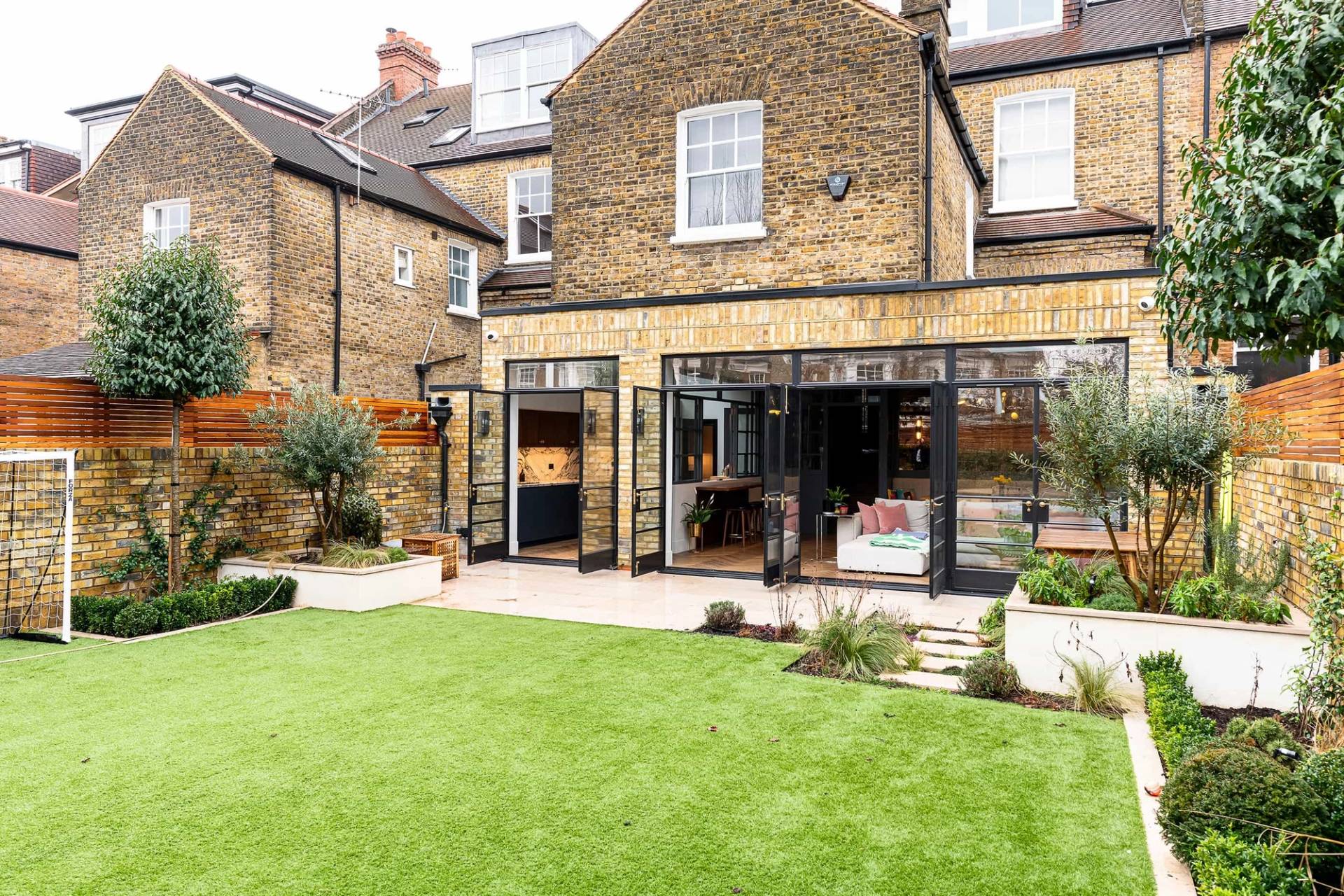 Rear exterior view of a yellow-brick London Victorian house showing a modern black crittall extension and manicured lawn, Love Locations.