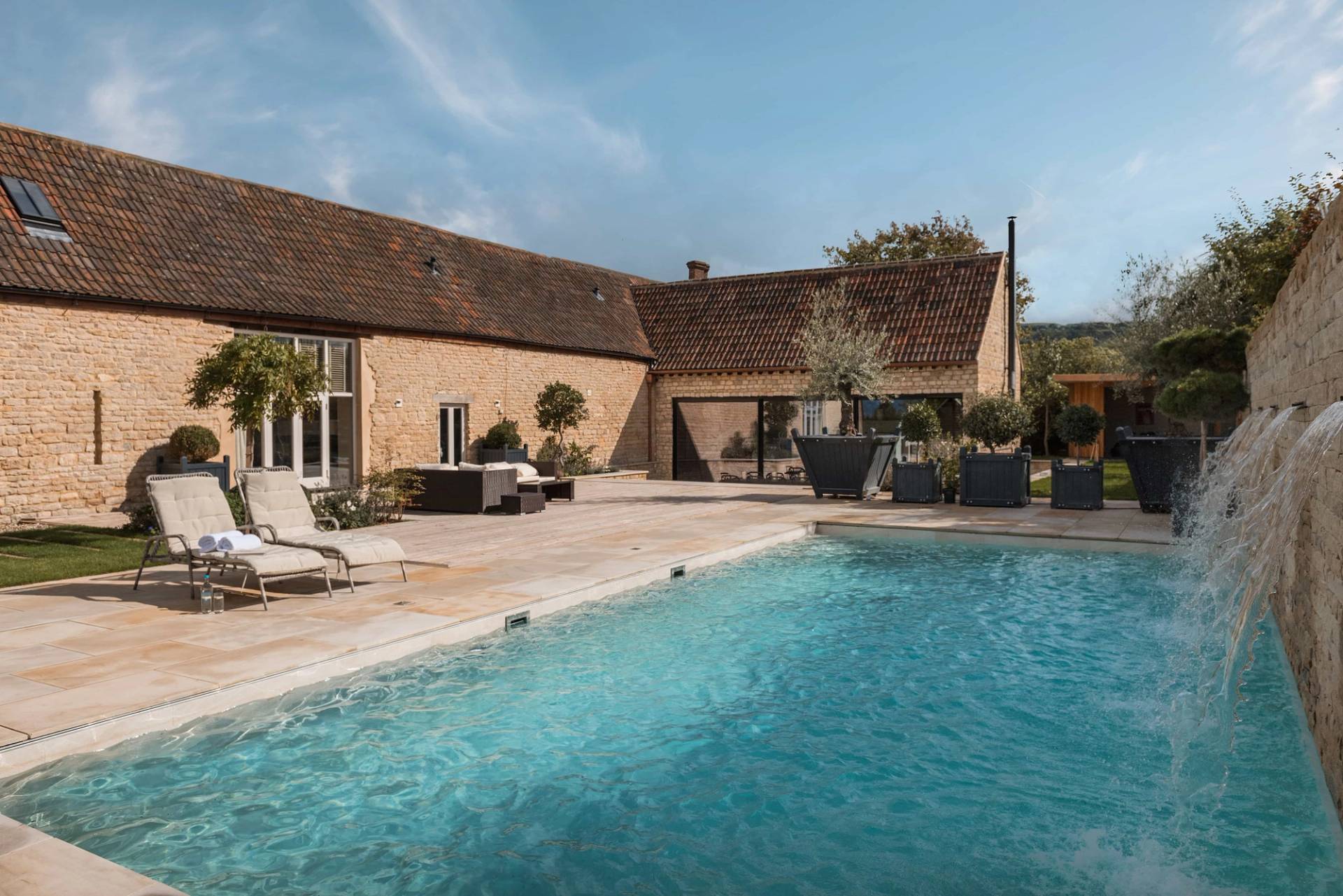 Luxury outdoor swimming pool terrace on a private Cotswold country estate in Gloucestershire GL51 through Love Locations, framed by stone outbuildings under a blue sky.