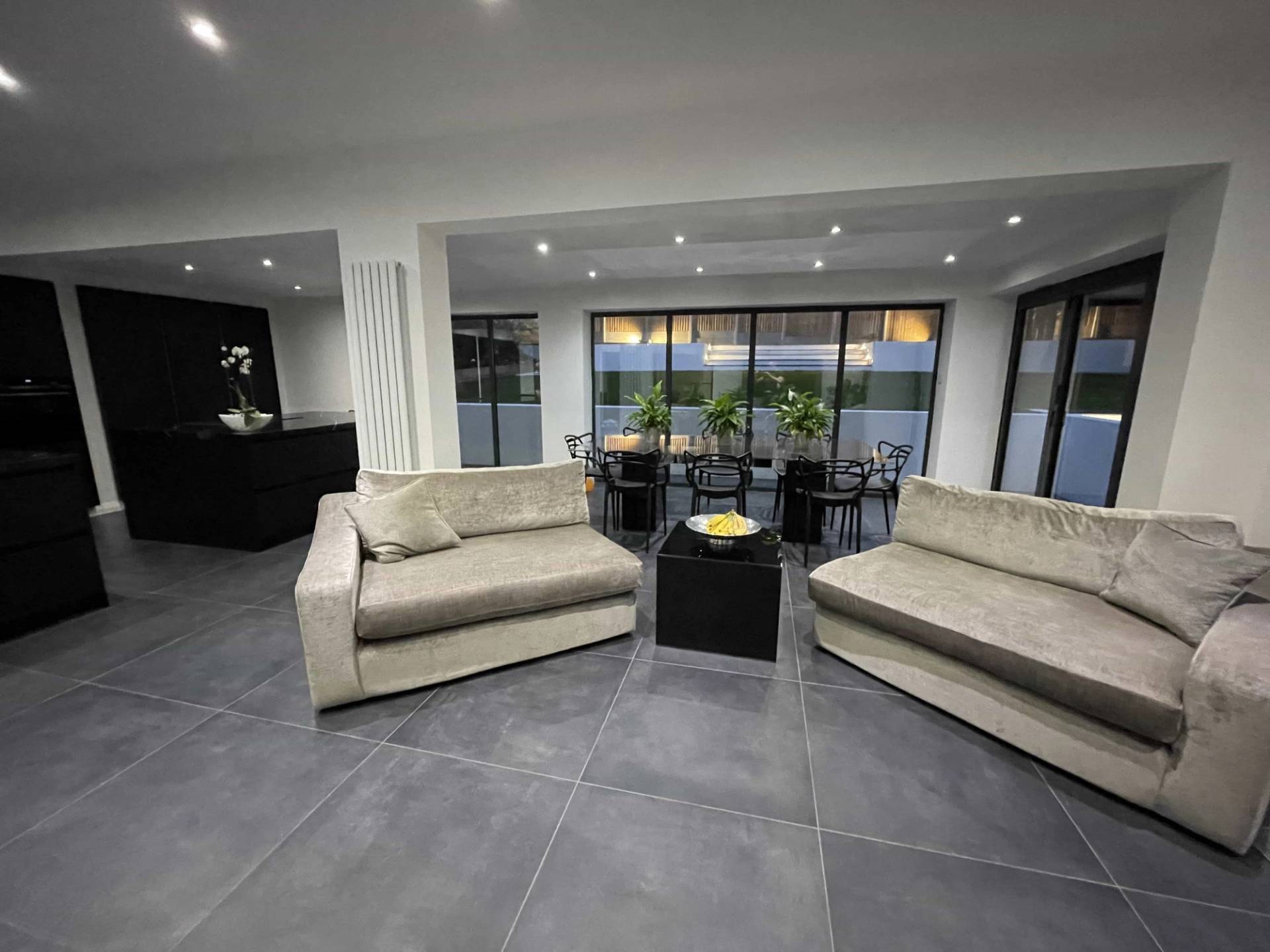 Glass garden room lounge featuring beige sofas, dark tiled floors, and full-height glass doors leading to a patio, Love Locations.