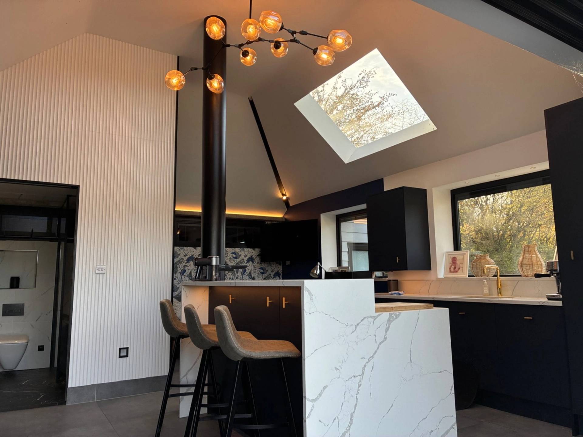 Modern vaulted ceiling pool house studio in Essex CM4 from Love Locations, displaying a white marble island, black bar stools, high ceilings, and skylights.