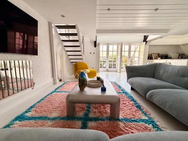 Sun-drenched open-plan penthouse living room inside a converted Victorian school in Brixton SW4 from Love Locations, featuring a colorful geometric rug, a cream sofa, and steps leading to a roof terrace.