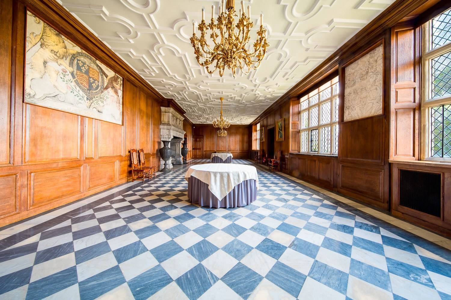 Historic oak-panelled Great Hall film location in Hertfordshire AL9 via Love Locations, featuring black-and-white chequered marble flooring, detailed decorative ceiling mouldings, and a brass chandelier.