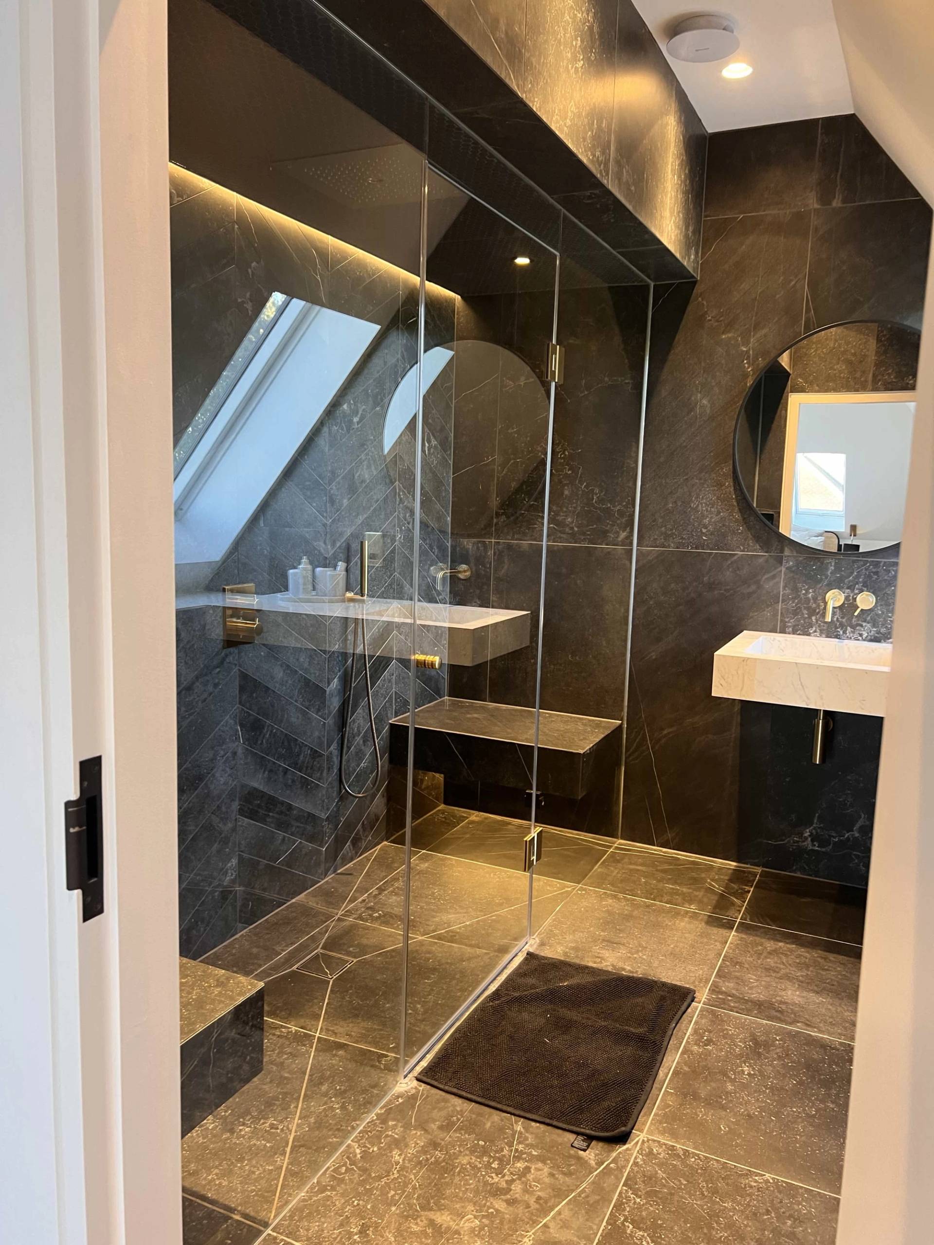 Modern dark marble shower room with a sloped ceiling skylight, walk-in glass screen, and recessed LED shelf lighting, Love Locations.