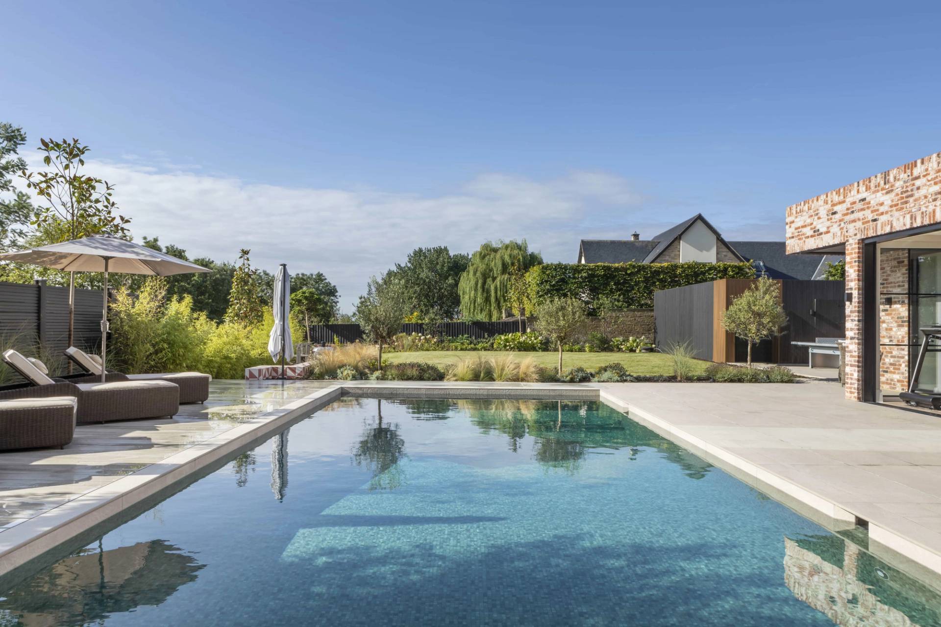 Clear blue swimming pool reflecting green trees and a modern brick country home in Buckinghamshire, Love Locations HP18.