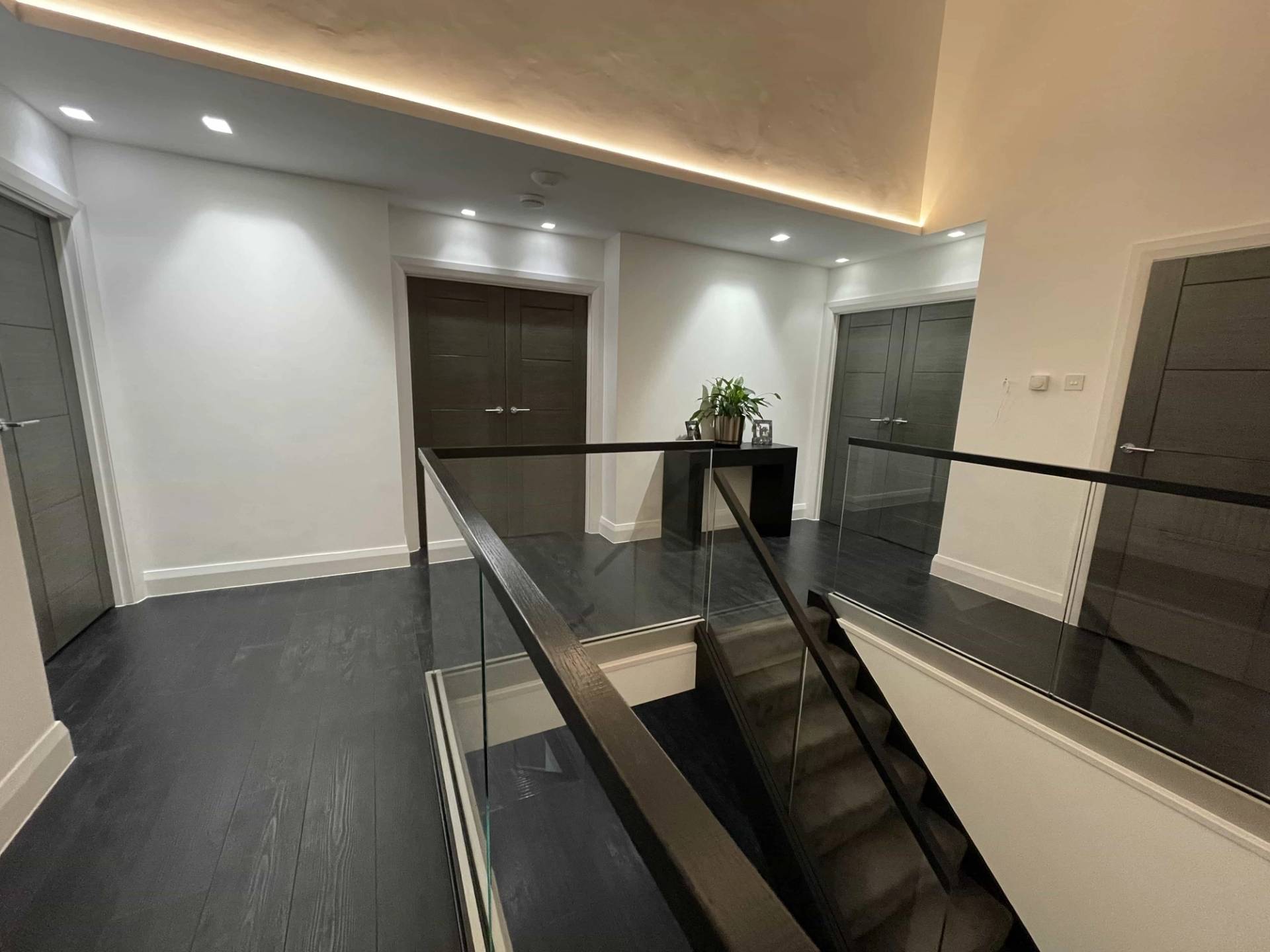 Double-height stairwell landing with dark timber flooring, glass balustrades, recessed doors, and soft ceiling trough LED lighting, ideal for fashion shoot transitions, Love Locations IG10.