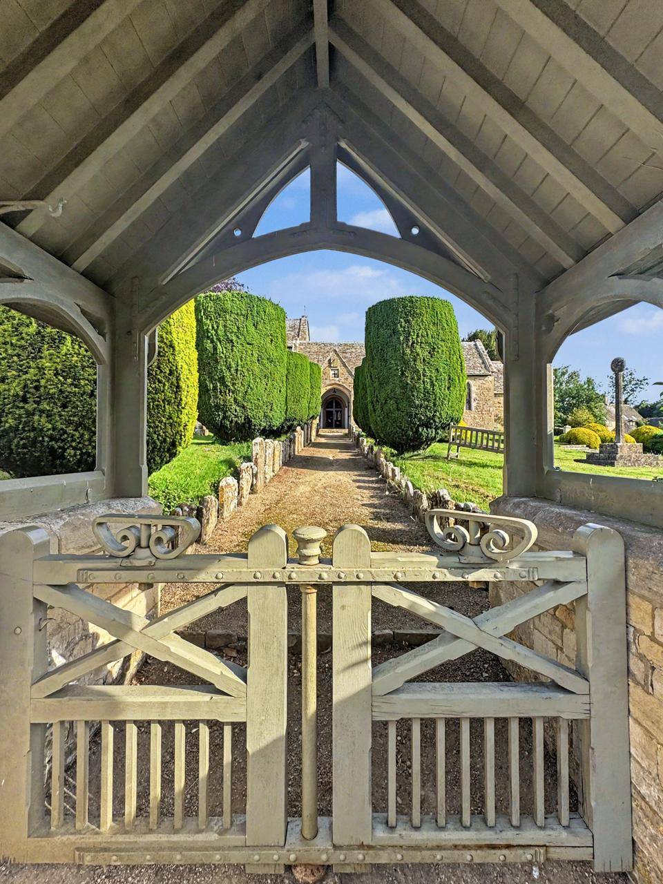 Underneath a wooden lychgate entry photoshoot location in Gloucestershire GL7 by Love Locations, looking down a gravel pathway through hedgerows toward the church.
