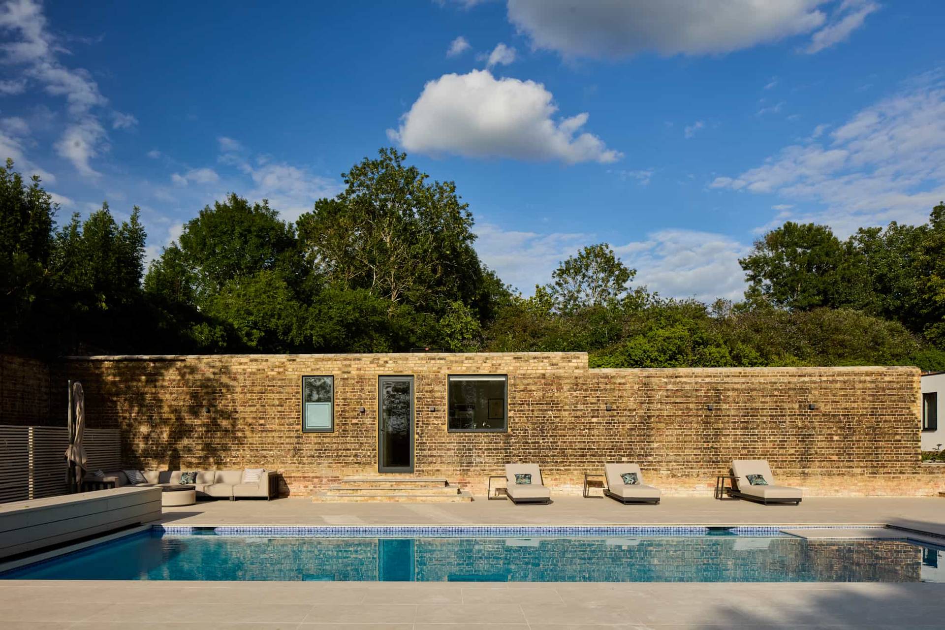 Wide-angle view of a large outdoor swimming pool and sun terrace set against a contemporary stone wall backdrop, Love Locations KT22.