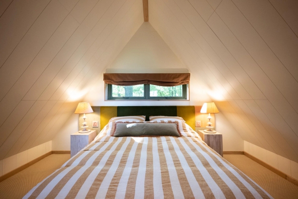 Symmetrical view of a double bed nestled under pointed exposed timber cabin rafters with warm bedside lighting, listed on Love Locations.