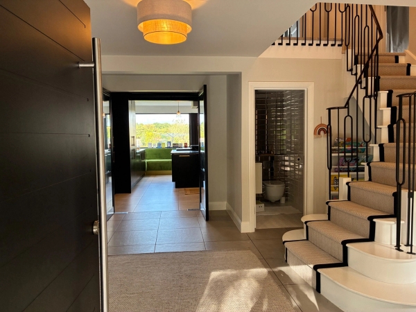 Bright grand foyer film location in Essex CM4 via Love Locations, showcasing a sweeping staircase, dark front door, wall sconces, and views out to the garden.