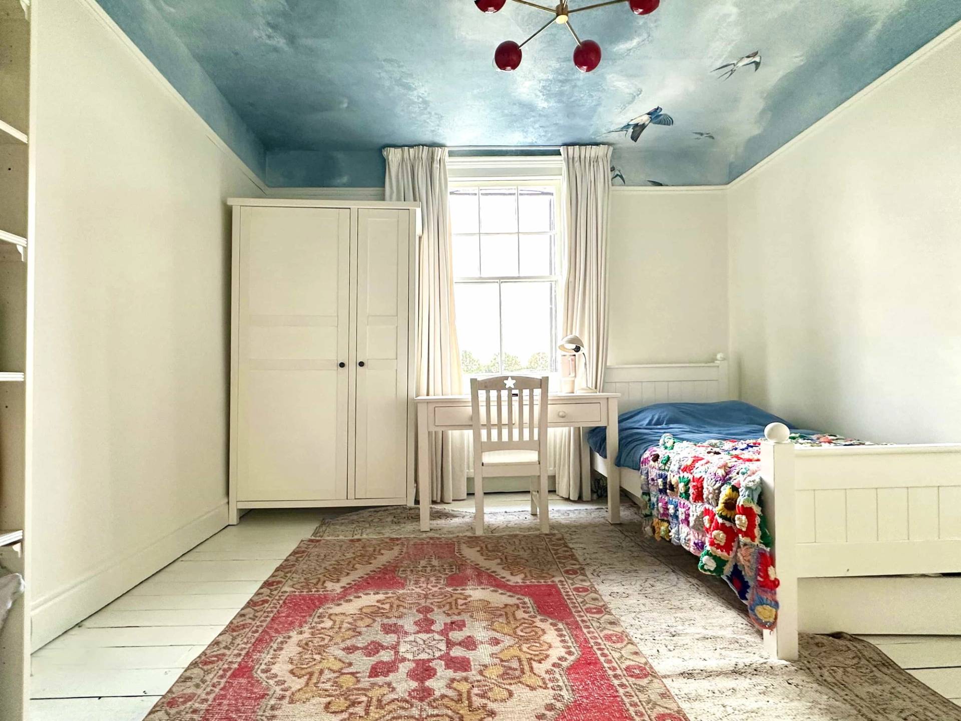 Eclectic bedroom with cloud-painted blue ceiling, white wardrobes, study desk, and red runner rug, Love Locations N13.