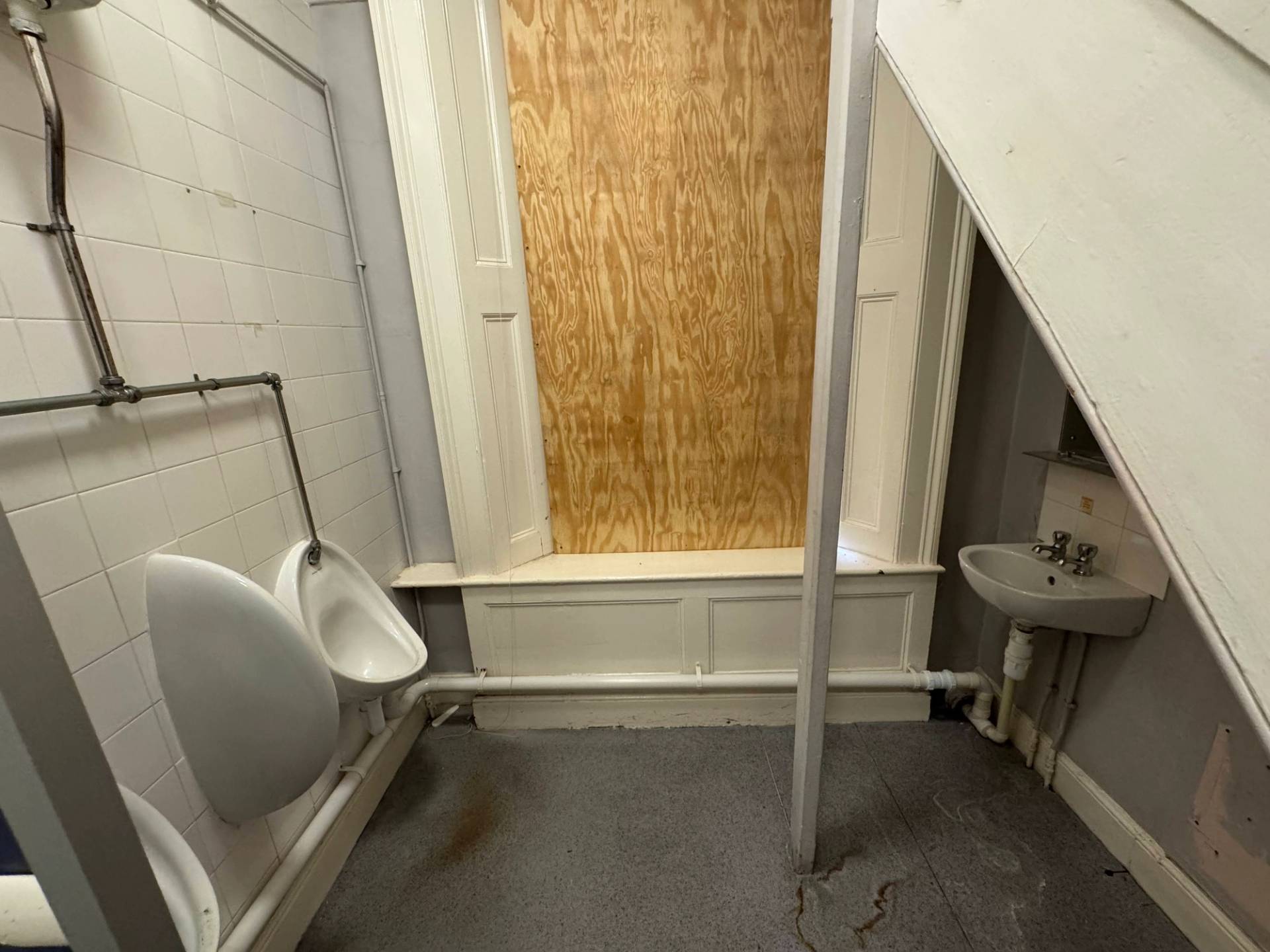 Dilapidated men's restroom showing wall-mounted urinals and a heavily boarded security window, Love Locations.