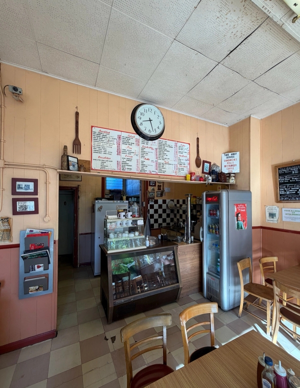 Traditional cafe counter area with menu chalkboard, glass display case, drinks fridge, and wall clock in Tulse Hill, Love Locations SE27.