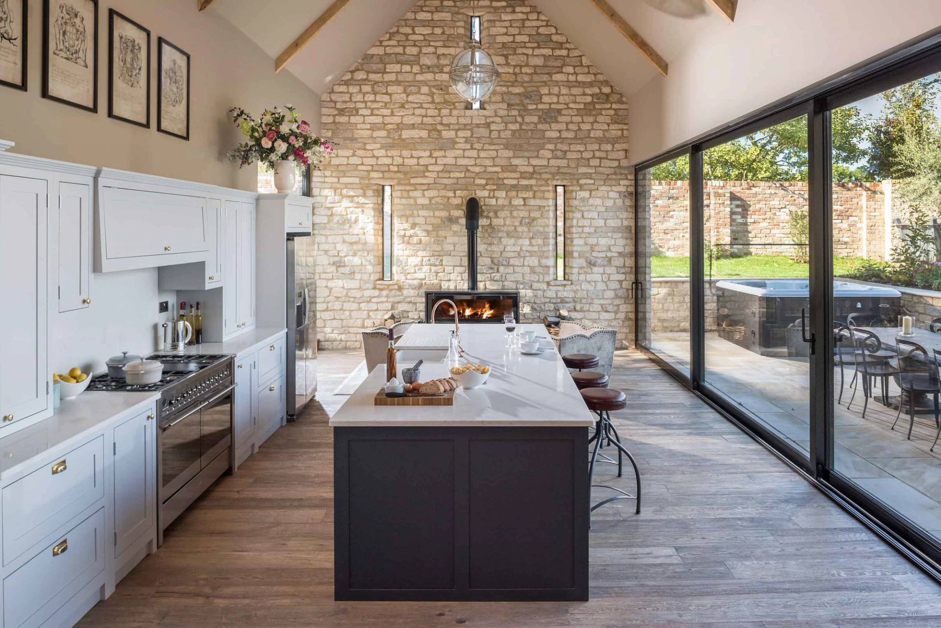 Vaulted open-plan country kitchen and dining location house in Cheltenham GL51 from Love Locations, featuring exposed Cotswold stone walls, a dark kitchen island, wood-burning stove, and glass bi-fold doors.