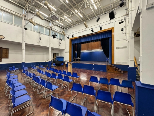 Love Locations secondary school hall shoot location Cockfosters EN4: Large assembly space featuring rows of blue chairs facing a theatrical wooden stage, ideal for school plays, TV dramas, and feature film productions.