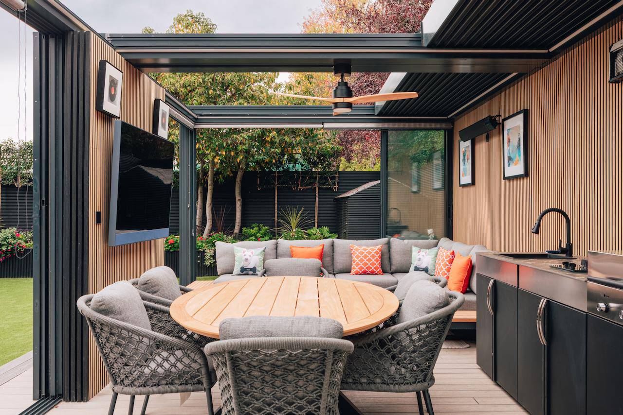 Enclosed outdoor pergola seating area featuring a round wooden dining table, woven chairs, ceiling fan, and outdoor TV, Love Locations.