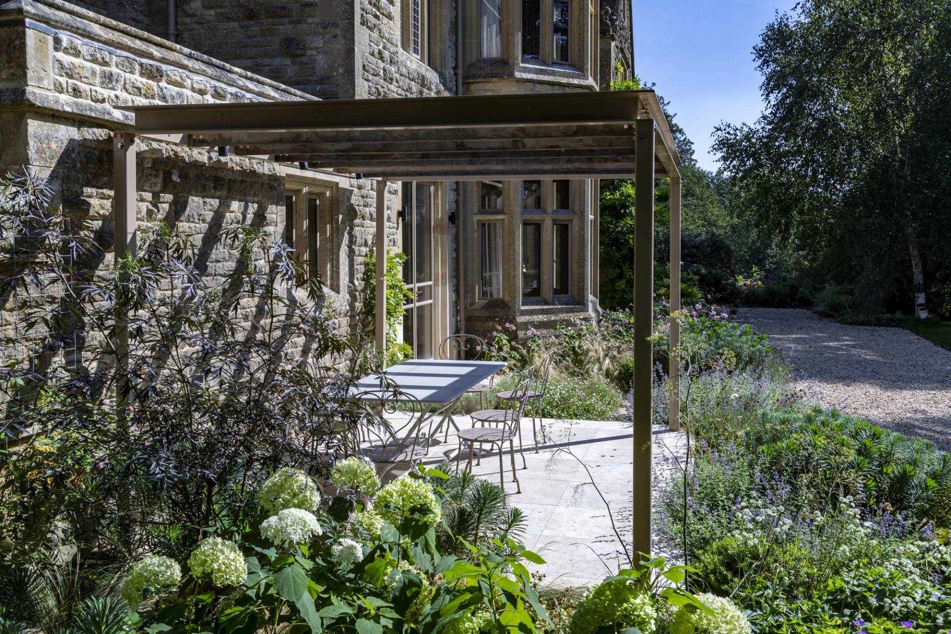 Paved outdoor terrace with hydrangeas set under a timber pergola outside stone house walls, Love Locations OX7.