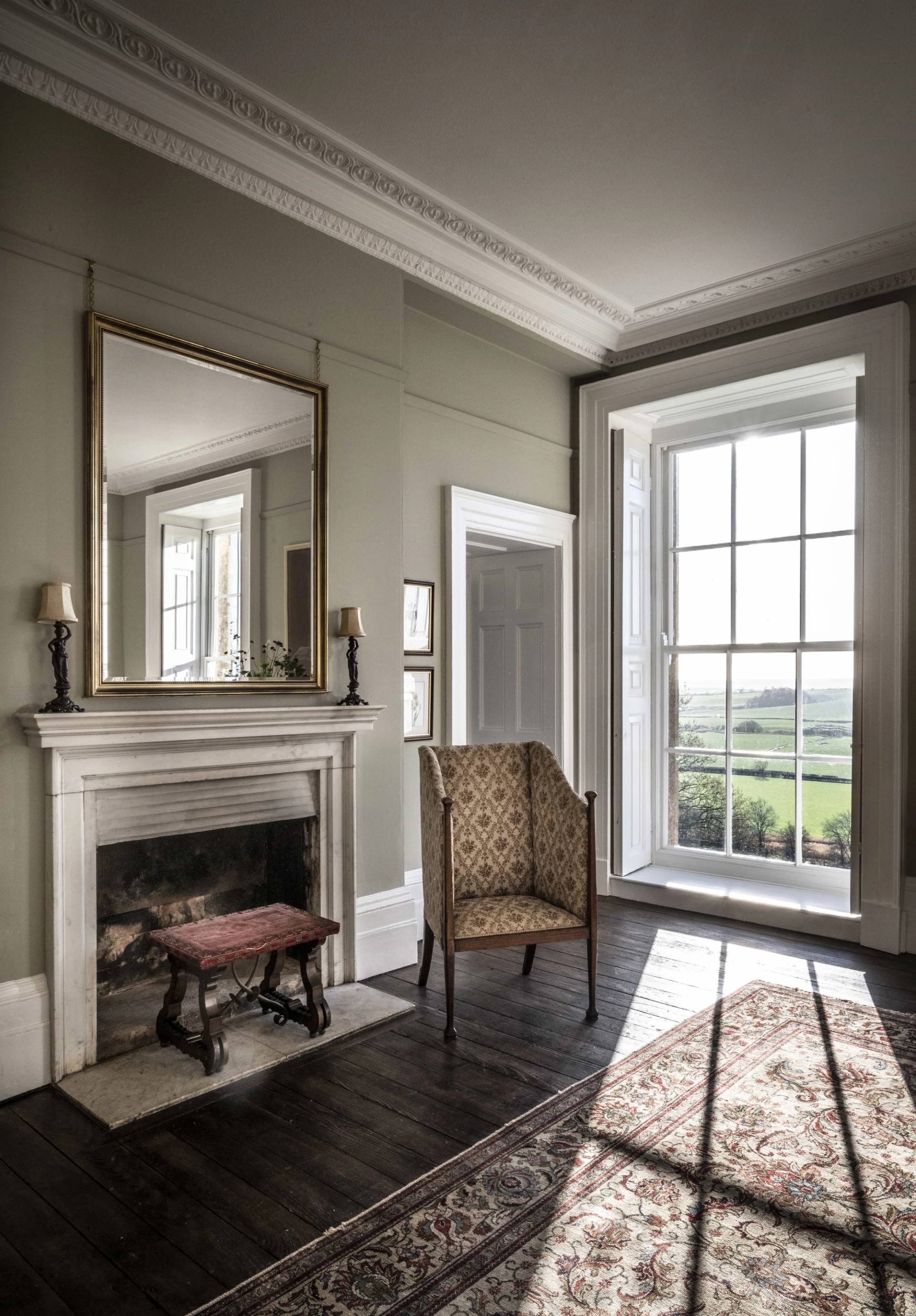 Sage green bedroom corner with a stone fireplace hearth, patterned armchair, and dark wooden floors, Love Locations.