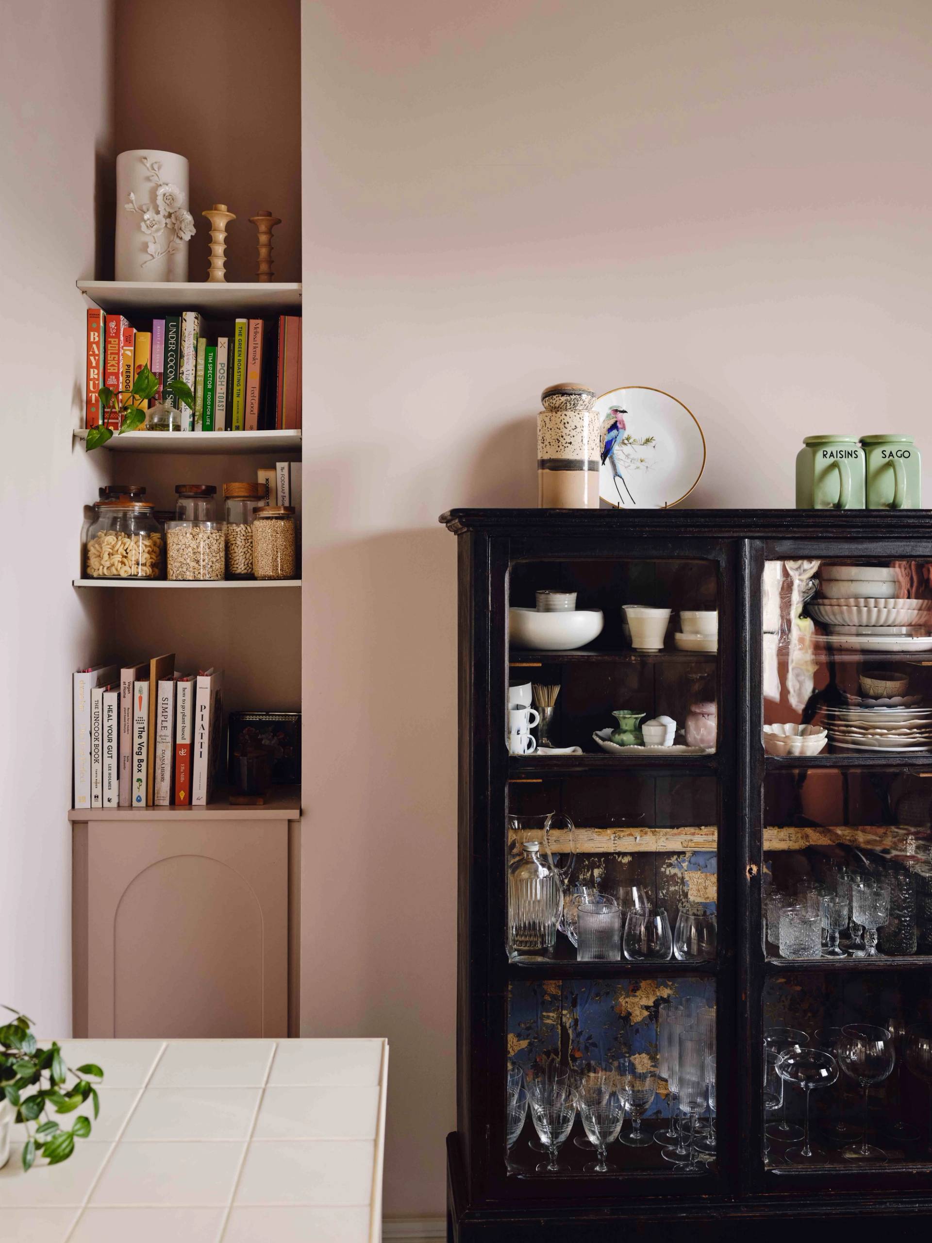 Dark vintage glass-front china cabinet set against plaster pink walls with open pantry shelving, Love Locations CR0.