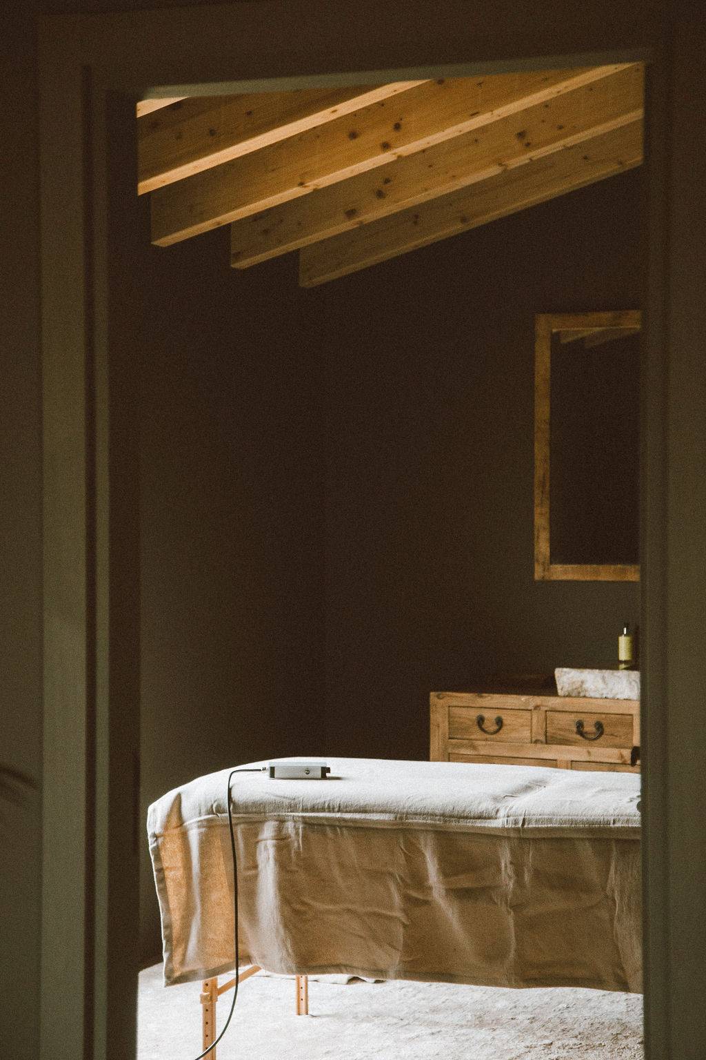 Spa massage room with a massage bed, dark green microcement walls, and wood ceiling beams, Love Locations 07630.