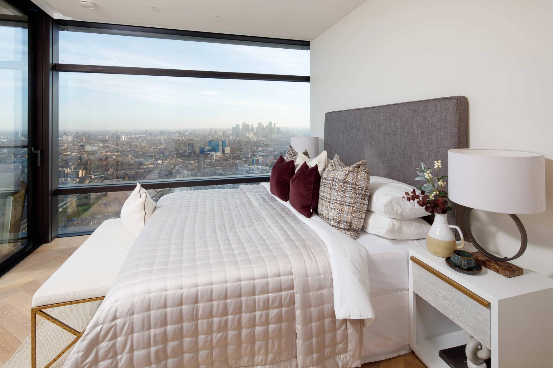 Luxury penthouse master bed layout styled with neutral quilted linens and burgundy velvet cushions overlooking East London, Love Locations EC2A.
