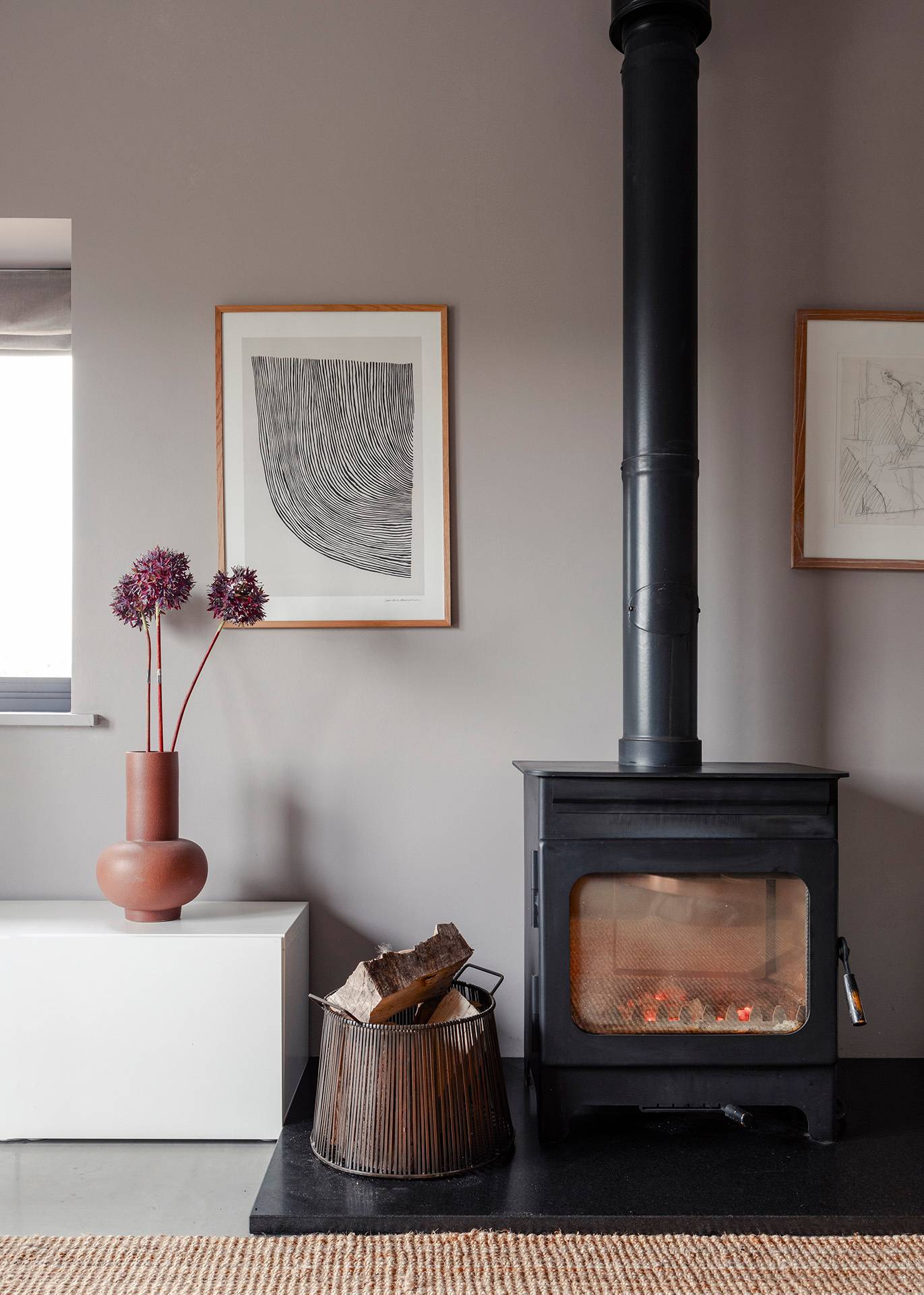 Minimalist wood-burning stove set against grey walls with a terracotta vase and abstract art print, Love Locations SN8.
