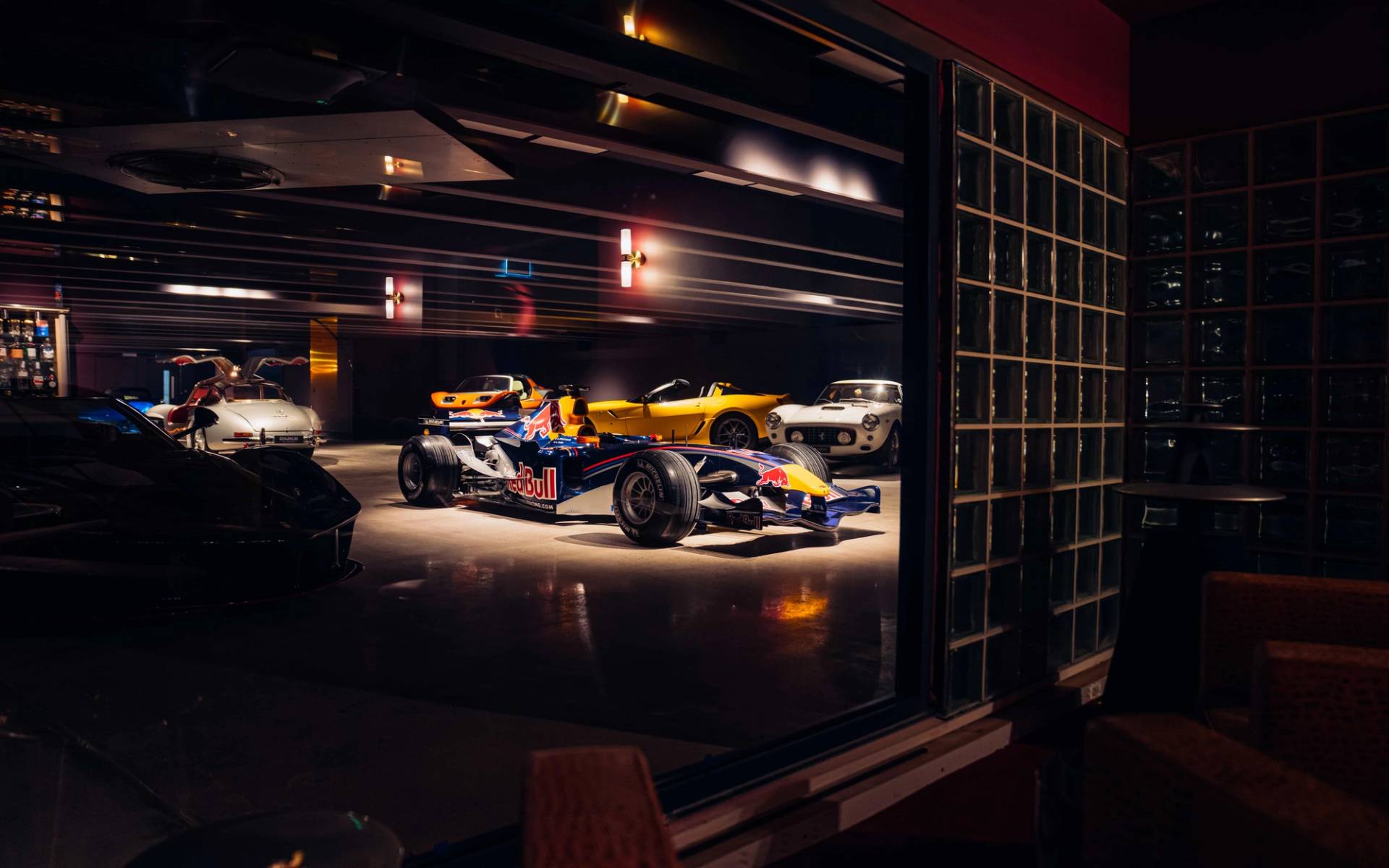 Nighttime glass window perspective into a dark underground supercar bunker displaying a Formula 1 car through Love Locations.