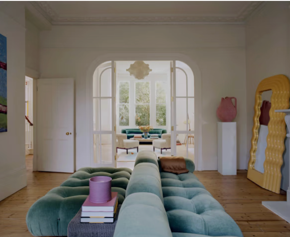 Open-plan living room featuring a tufted green velvet sofa, large yellow wavy mirror, sculptural ceramics, timber floors, and an arched opening leading to a light-filled sitting room with tall sash windows.