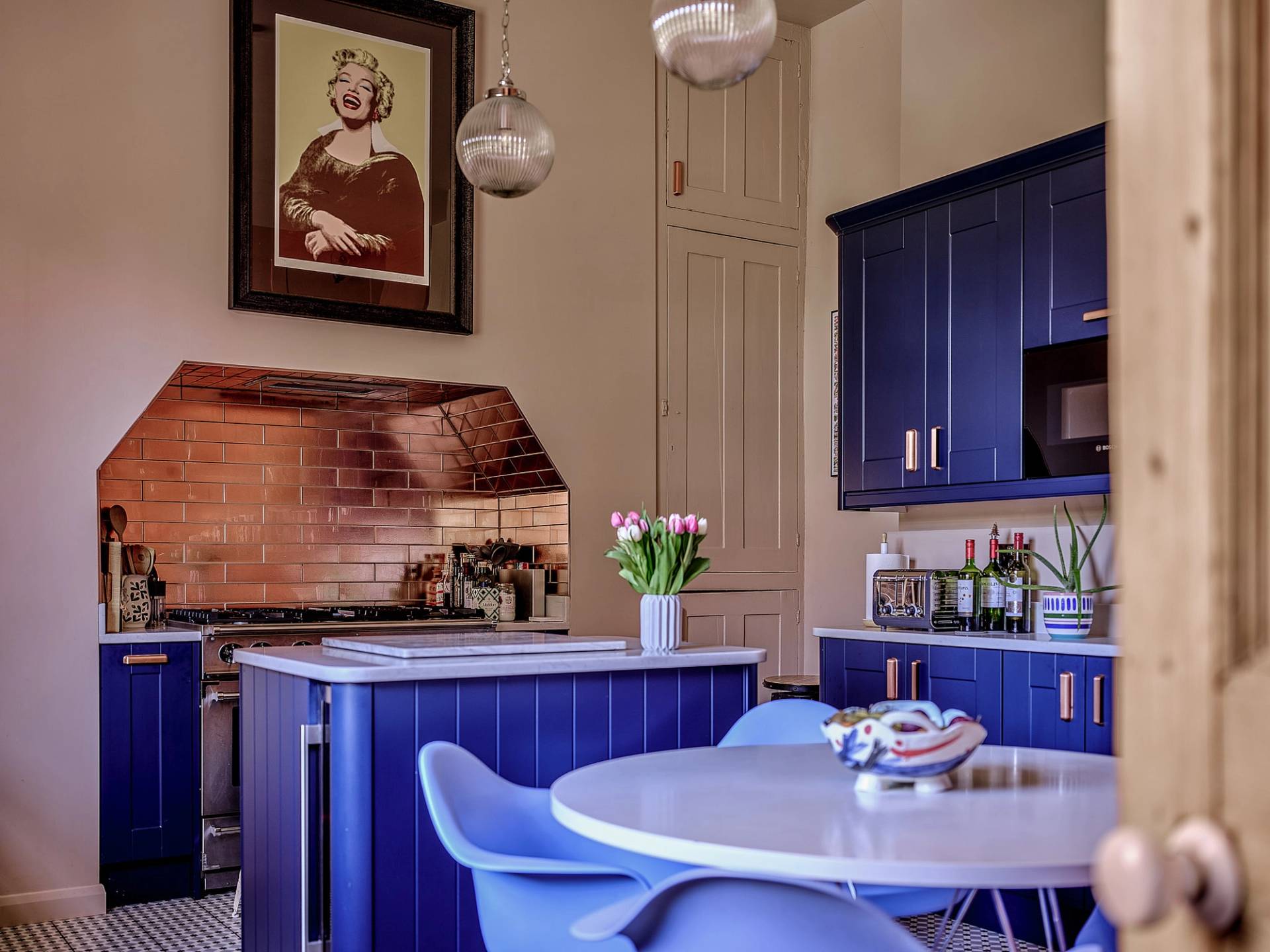 Eclectic kitchen featuring cobalt blue island, white dining table, blue chairs, and copper extractor hood detail, Love Locations.