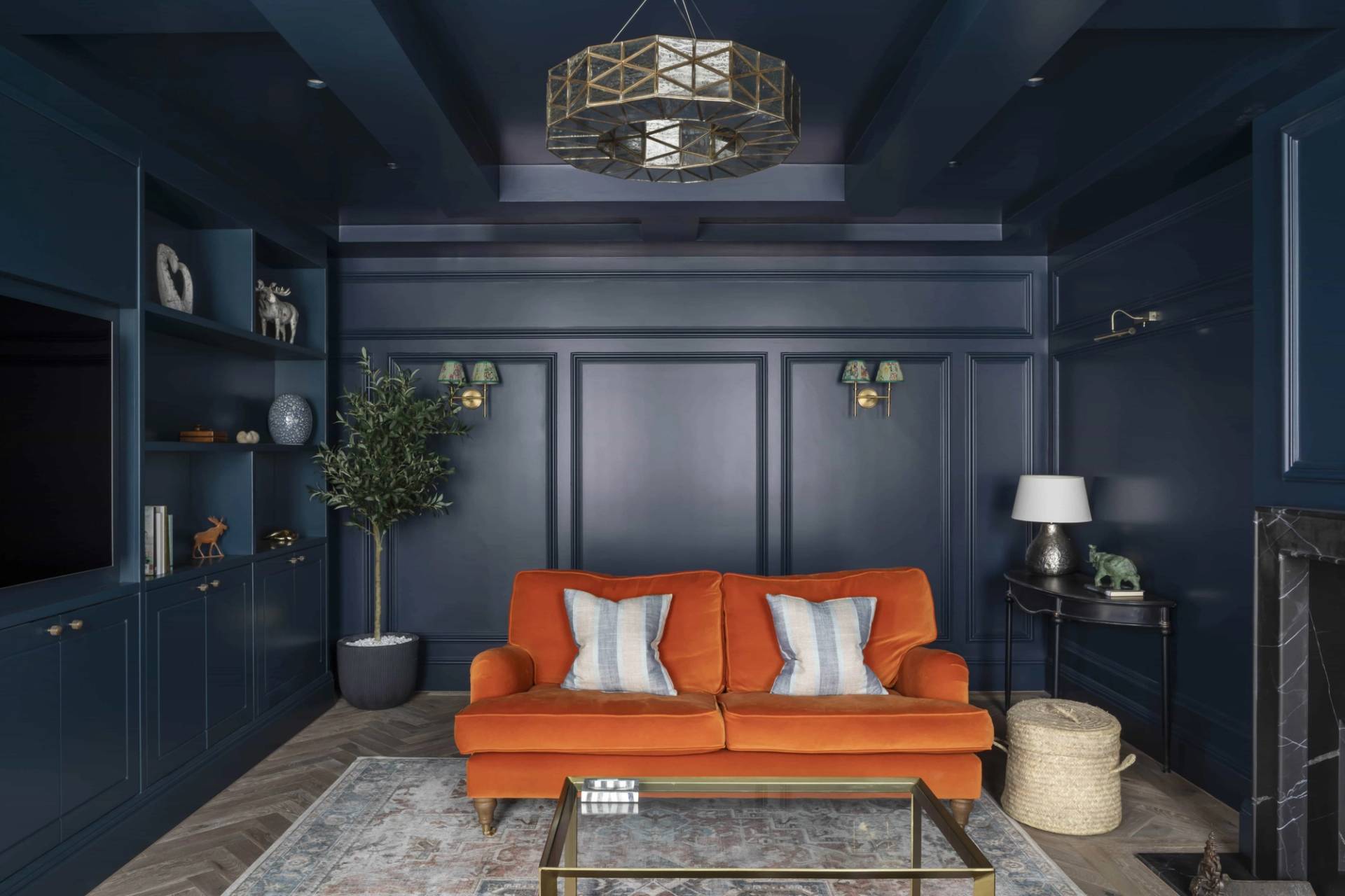 Symmetrical shot of a dark navy panelled lounge with a vibrant orange velvet sofa and brass sconces, Love Locations HP18.